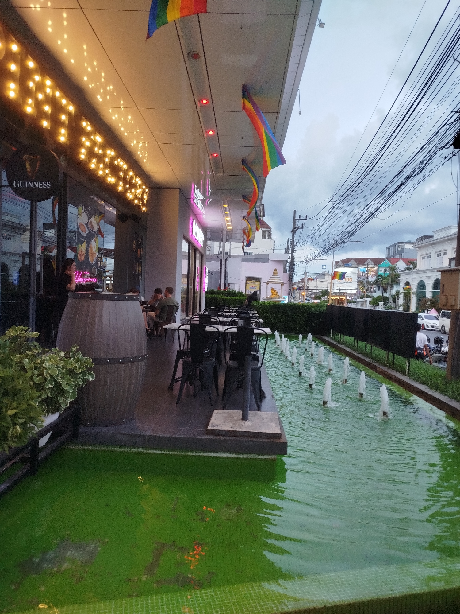 A wide shot captures an outdoor seating area of a establishment with outdoor lighting and several rainbow flags. In the foreground, a green, grassy area is visible with a tiled border. A water feature with multiple spouts runs parallel to the seating area, which includes tables and chairs. Behind the seating area, a building facade is visible, featuring a Guinness sign and illuminated signage. People are seated at tables, and a server is standing behind a wooden barrel. In the background, a street scene unfolds with buildings, vehicles, and a prominent rainbow flag displayed on a building. The sky is overcast, and power lines are strung across the upper portion of the image.