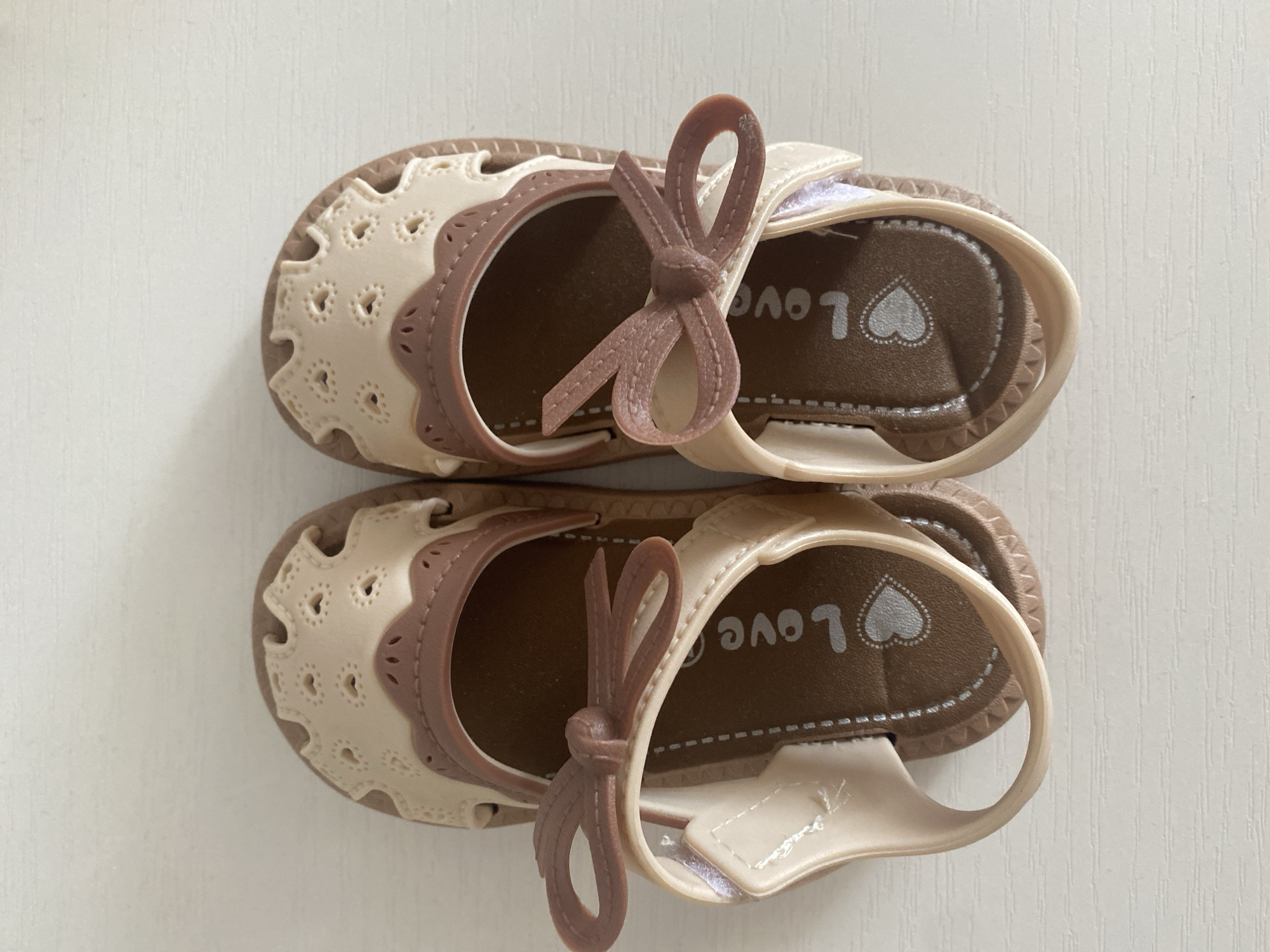 A pair of light beige and brown children's sandals are displayed from a top-down perspective on a white, textured surface. The sandals have decorative cutouts on the front, resembling hearts, and are adorned with small, light brown bows on each strap. The insoles are brown and feature the word "Love" and a heart design. The background is a plain white surface with subtle horizontal lines. No people are present in the image, and the location is not discernible.
