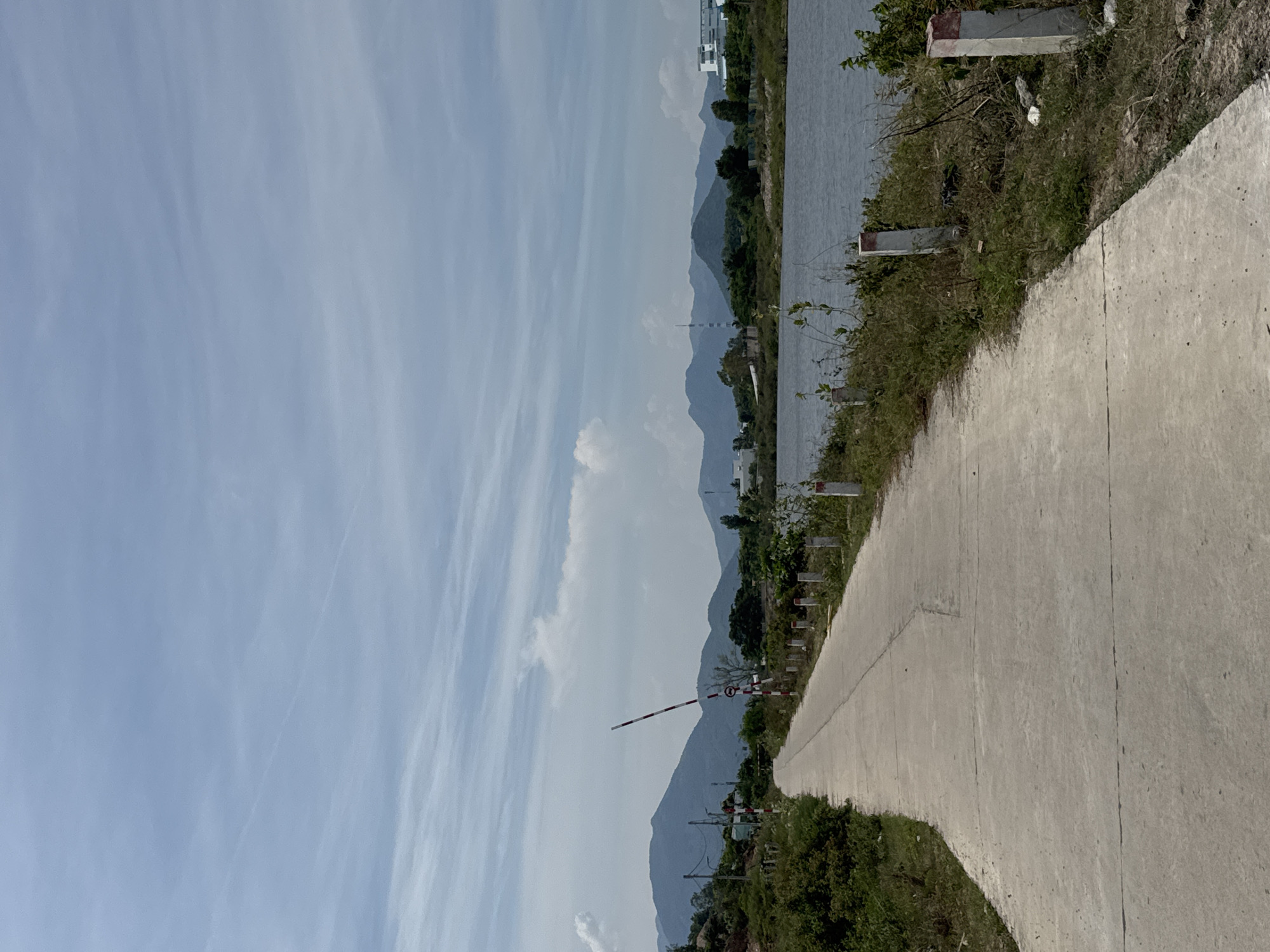 A paved road curves to the left, bordered by a body of water on the right and lush green vegetation on the left. In the distance, a range of misty blue mountains forms the horizon under a cloudy sky. A red and white striped barrier gate stands on the left side of the road, near a traffic sign. Several concrete posts with red tops mark the edge of the road near the water. The environment appears to be a rural or semi-rural area in Nam Nha Trang Ward, Vietnam. There are no people visible in the image.