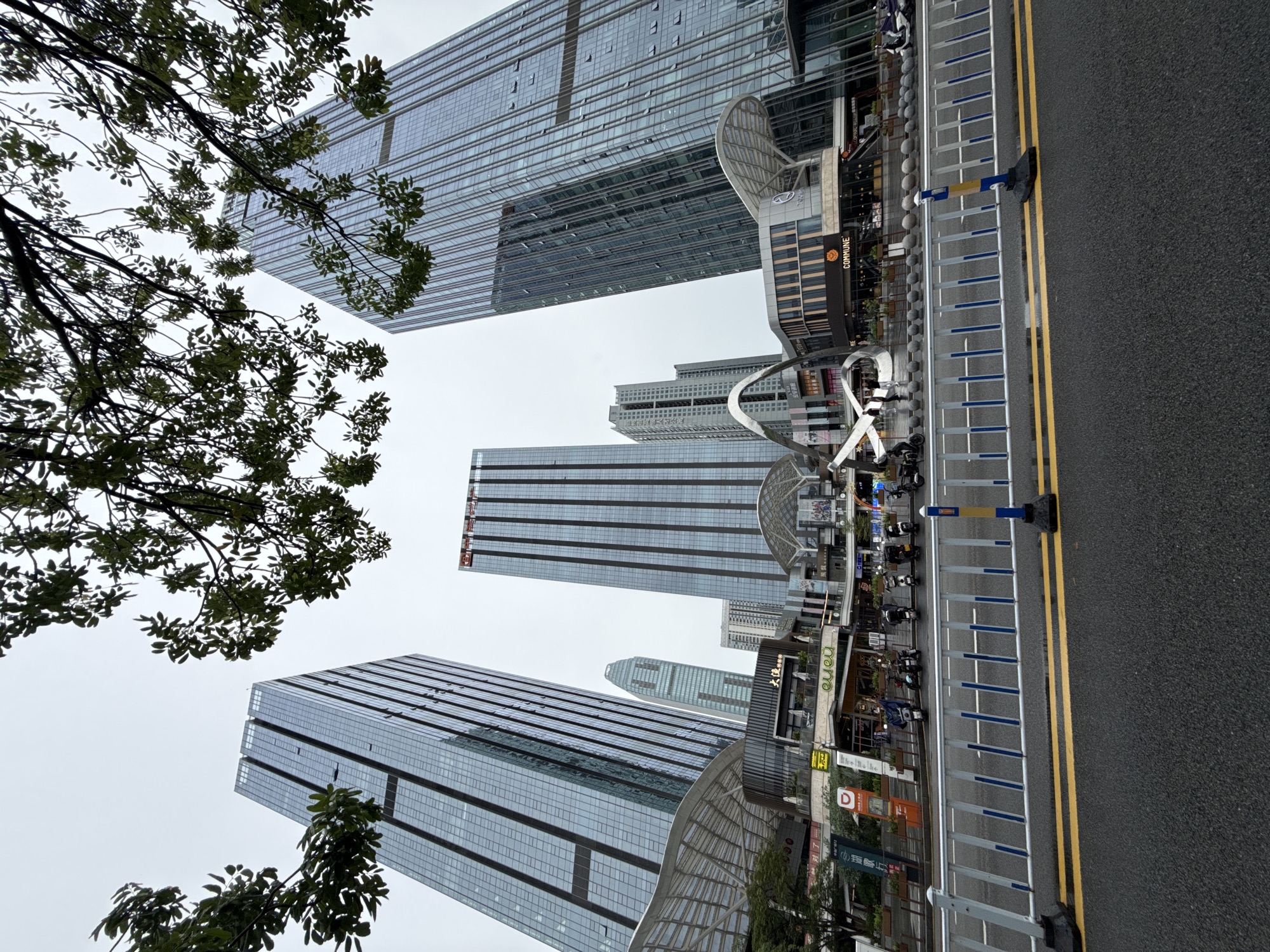 The image depicts a wide-angle view of a modern urban street scene in Huizhou, China, with a focus on contemporary architecture and commercial spaces. In the foreground, a paved road with double yellow lines is visible, bordered by a white and blue striped barrier. Beyond the barrier, a row of parked motorcycles lines the sidewalk.
The background is dominated by several tall, glass-clad skyscrapers, reflecting the overcast sky. One building prominently displays Chinese characters and a red logo near its top. In front of the skyscrapers, a multi-story commercial complex is situated, featuring various storefronts and covered walkways with arched, translucent roofs. One storefront has the word "COMMUNE" clearly visible on its facade. Several trees with dark green foliage are positioned in the upper portion of the frame, partially obscuring the sky and buildings, suggesting an outdoor, possibly slightly damp, environment. The overall impression is one of a bustling commercial district within a contemporary Chinese city.