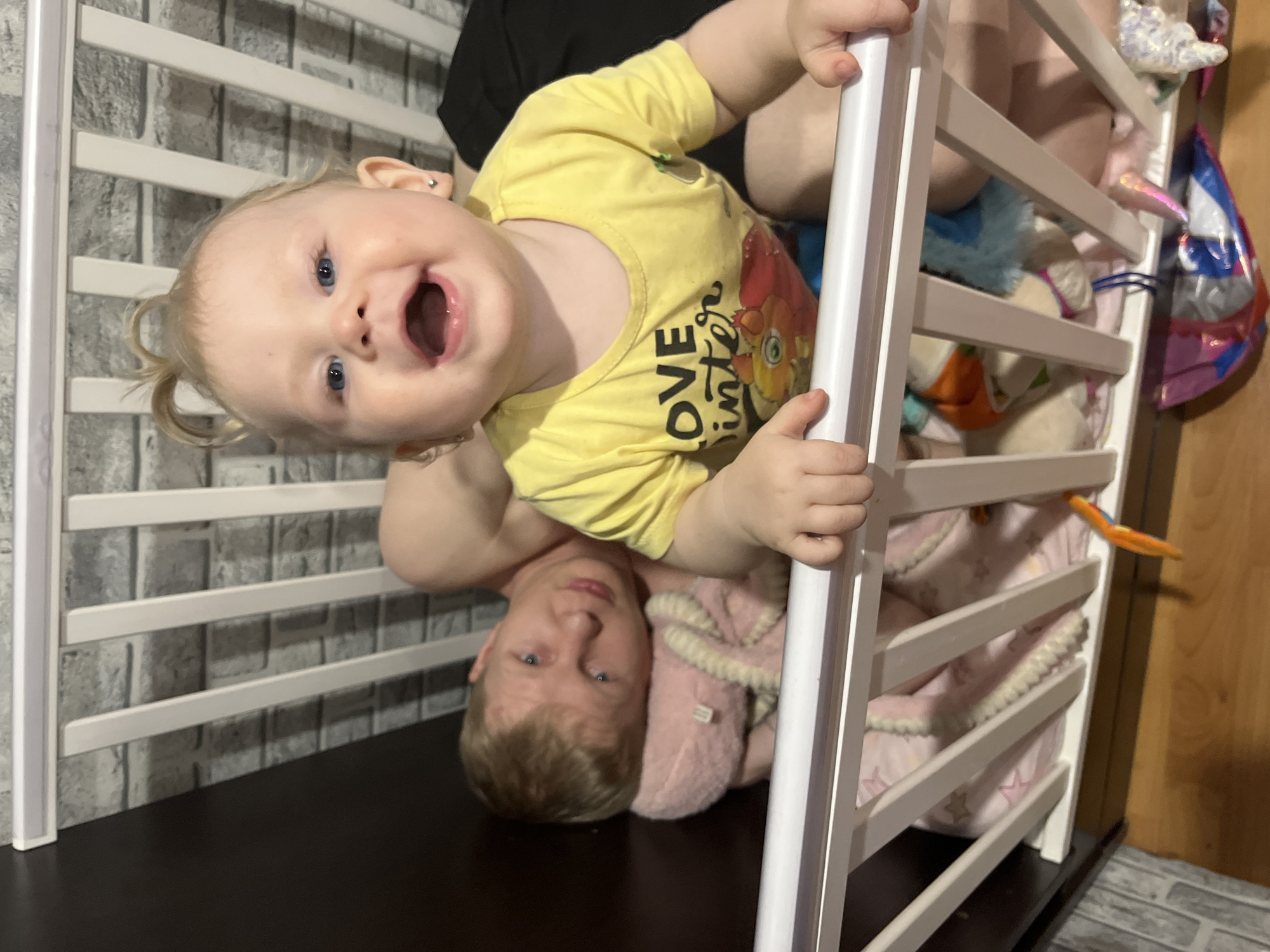 A photograph, taken indoors, shows a Caucasian baby in a yellow shirt, suspended upside down from a white crib. An adult is underneath the child. The baby has fair skin, blonde pigtails, and blue eyes. The adult is fair-skinned with short blonde hair. The crib contains various plush toys and pink blankets with white stars. A dark-colored wall and flooring are visible in the background. The location is Vesyoloye, Russia.