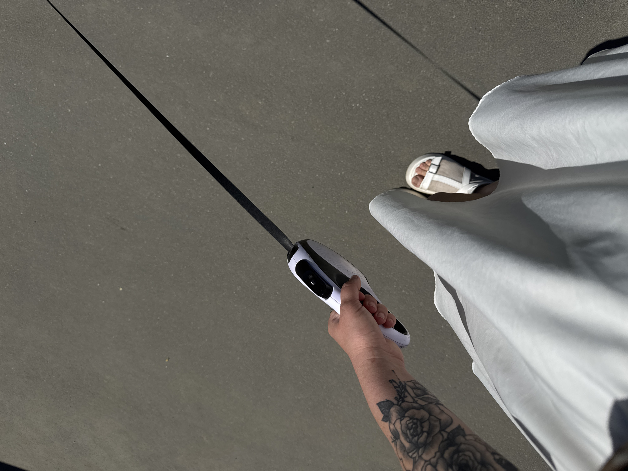 A first-person, high-angle shot shows an arm with a floral tattoo holding a white and black electric vehicle charging connector, with the attached black charging cable stretching diagonally upward to the left. A woman wearing a long white dress and white sandals stands on asphalt paving, part of an urban street. The location is Unknown, Unknown.