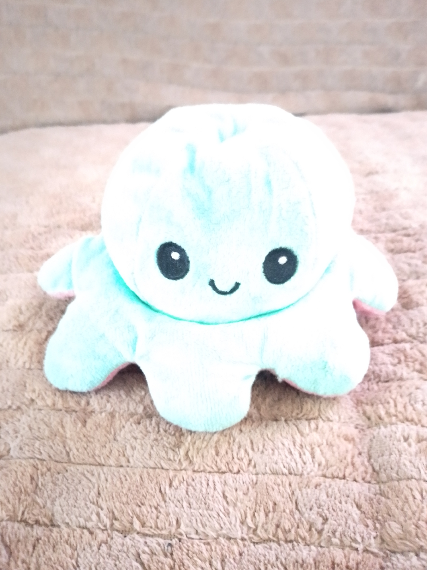 The image shows a close-up, eye-level shot of a reversible plush toy octopus. The octopus is light blue on one side and pink on the other, with its blue side currently displayed. It has two black embroidered eyes and a small, curved black embroidered smile. The octopus is lying on a textured, light brown blanket or rug, which has a ribbed or corduroy-like pattern. The lighting is soft and diffused, suggesting it might be indoors or on an overcast day. There are no people visible in the image. The focus is solely on the plush toy, with the background slightly out of focus. The overall mood conveyed by the toy's expression is happy and friendly.