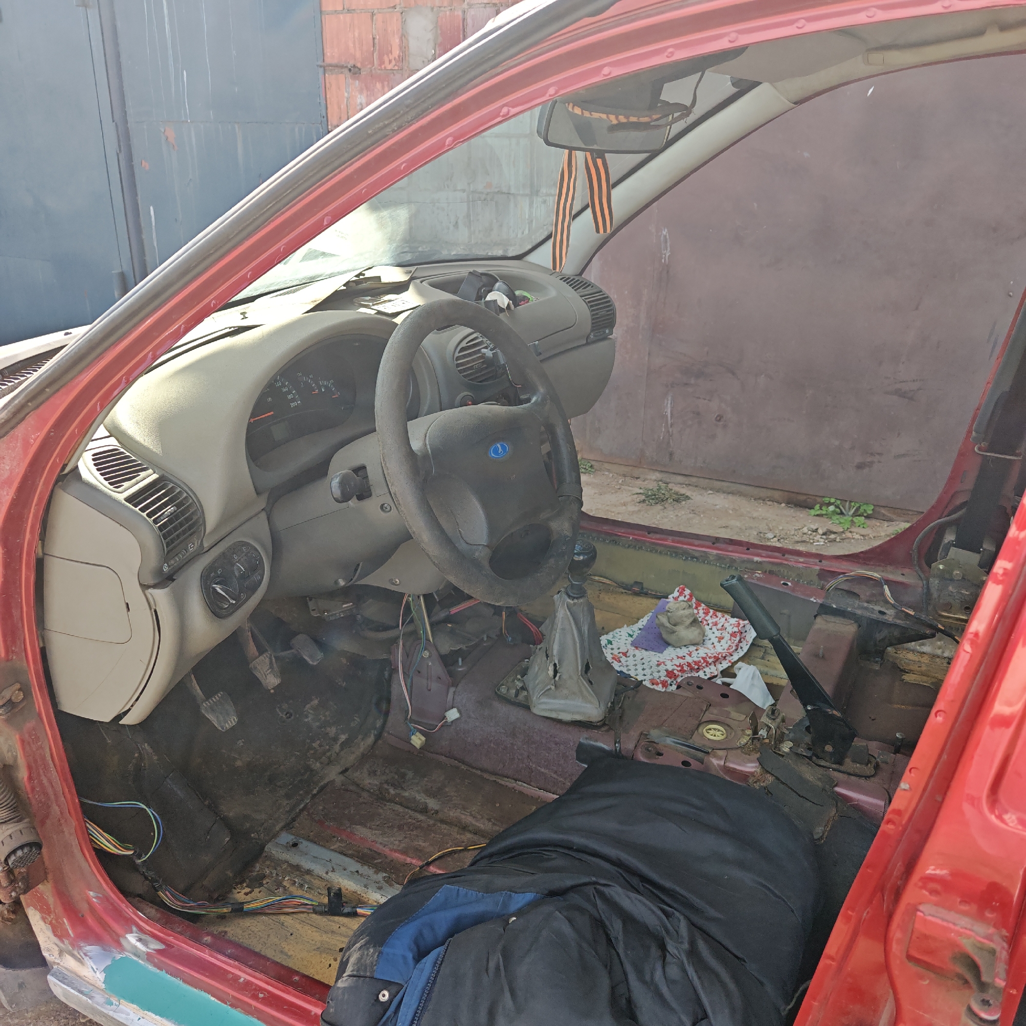 The interior of a disassembled red car is visible, likely a Lada Kalina, based on the steering wheel logo. The dashboard is still in place, but the flooring, seats, and door panels have been removed, exposing the bare metal frame and wiring. A black jacket is lying on the floor. A St. George ribbon hangs from the rearview mirror. The scene appears to be in a garage or workshop setting, possibly in the city of Rostov, Russia, based on the potential car model.