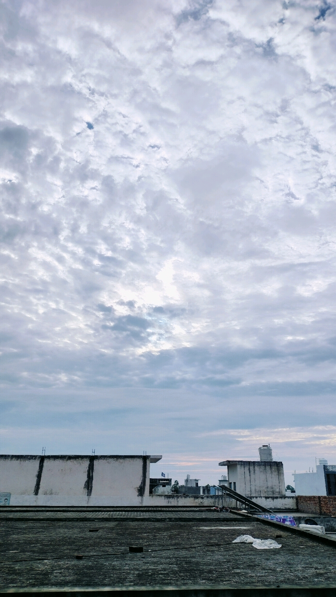A low-angle, vertical shot captures a cloudy sky over the rooftops of Ramraj, India. The sky is filled with a dense layer of cumulonimbus clouds, ranging in color from white to dark gray, with patches of sunlight breaking through. Below the sky, several flat rooftops are visible. The closest rooftop in the foreground is made of dark, weathered material and shows scattered bricks. Further back, a series of white and gray buildings with flat roofs are visible. One building on the right has a large water tank on its roof. Some of the buildings show signs of wear and tear, with stained walls. Solar panels are visible on the roof of one building, angled towards the sky. The overall scene depicts an urban landscape under an overcast sky.