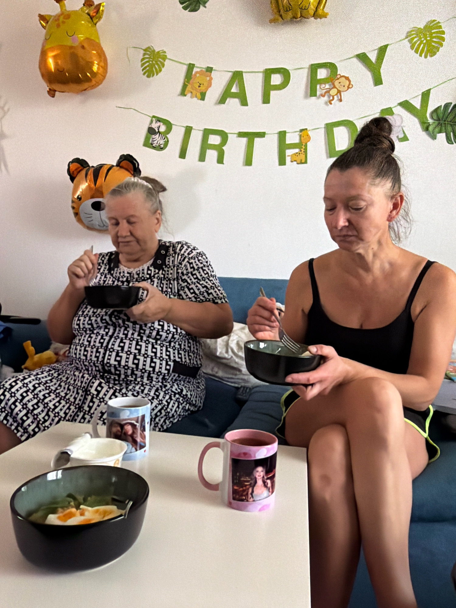 The image depicts a domestic indoor scene, likely a living room or dining area, with decorations suggesting a birthday celebration. Two women are seated on a blue sofa, each holding a bowl and a fork, engaged in eating. The woman on the left, who appears older, wears a black and white patterned dress. The woman on the right, with her hair in a bun, is dressed in a black tank top and shorts. In the background, a white wall is adorned with a "HAPPY BIRTHDAY" banner in green letters, featuring jungle-themed animal cutouts and leaves. Two foil balloons, shaped like a giraffe and a tiger's head, are also visible on the wall. A white table in front of the sofa holds several items, including bowls of food, mugs with personal photos, and a napkin. The lighting suggests an indoor environment, possibly during the daytime. There are no elements in the image that specifically indicate the location is Sharm El Sheikh, Egypt; the scene is generic to many indoor domestic settings.