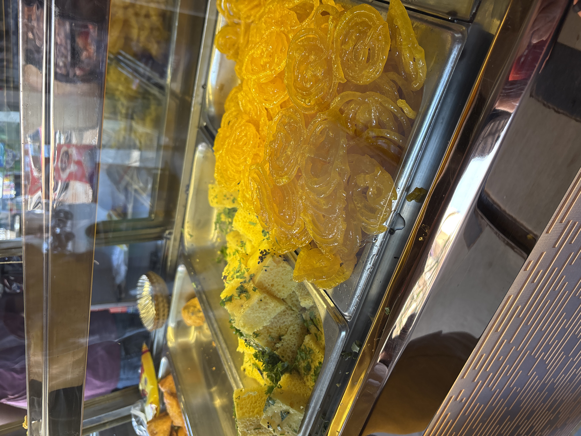 The image shows a display case in Ambernath, India, filled with Indian sweets. The top tray contains jalebi, a bright yellow, spiral-shaped sweet. Below the jalebi is a tray of dhokla, a spongy, yellow, savory snack garnished with cilantro and mustard seeds. The sweets are visible through the glass front of the display case. Other food items are partially visible in the trays to the left. The setting appears to be a food shop.