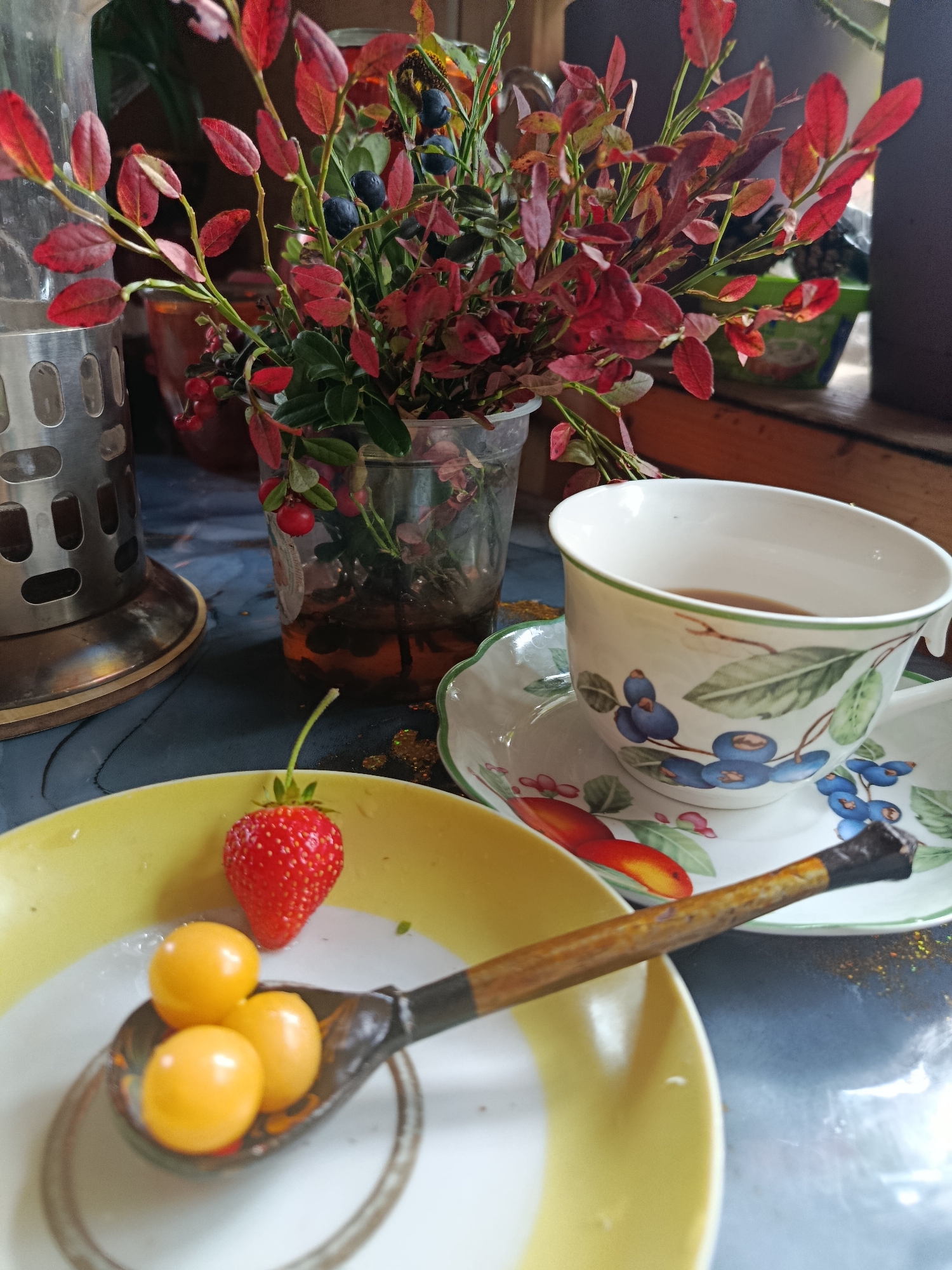 A close-up shot captures a table setting with a rustic and natural aesthetic. In the foreground, a yellow-rimmed plate holds a single ripe strawberry and a wooden spoon filled with three small, round, yellow fruits. Behind this, a white teacup and saucer, adorned with a pattern of blueberries and leaves, rests on the table. The teacup contains a dark liquid, presumably tea. To the left, a metallic cylindrical object, possibly a tea infuser or a decorative holder, stands prominently. In the background, a bouquet of branches with vibrant red leaves and dark blue berries is arranged in a clear container, adding a touch of autumnal color and woodland charm to the scene. The overall impression is one of a simple, yet inviting, moment of enjoying nature's bounty.