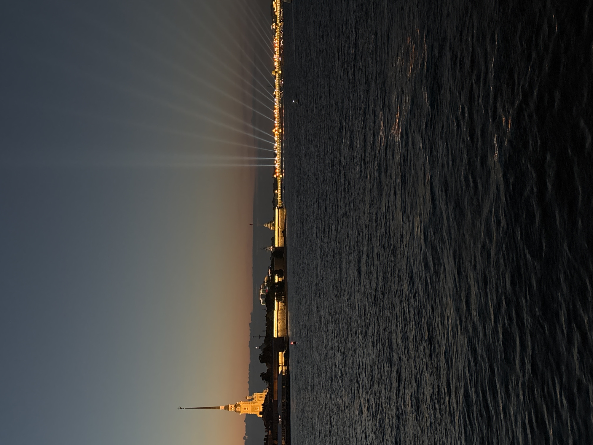 The image depicts a nighttime view of the Peter and Paul Fortress in Saint Petersburg, Russia, across the Neva River. The fortress is illuminated, with its iconic spire and buildings clearly visible against the dark sky. A series of bright spotlights emanate from the riverbank, casting dramatic beams upwards into the sky. The river itself is dark and choppy, reflecting the ambient light from the city. The overall scene conveys a sense of grandeur and nighttime illumination of a significant historical landmark.