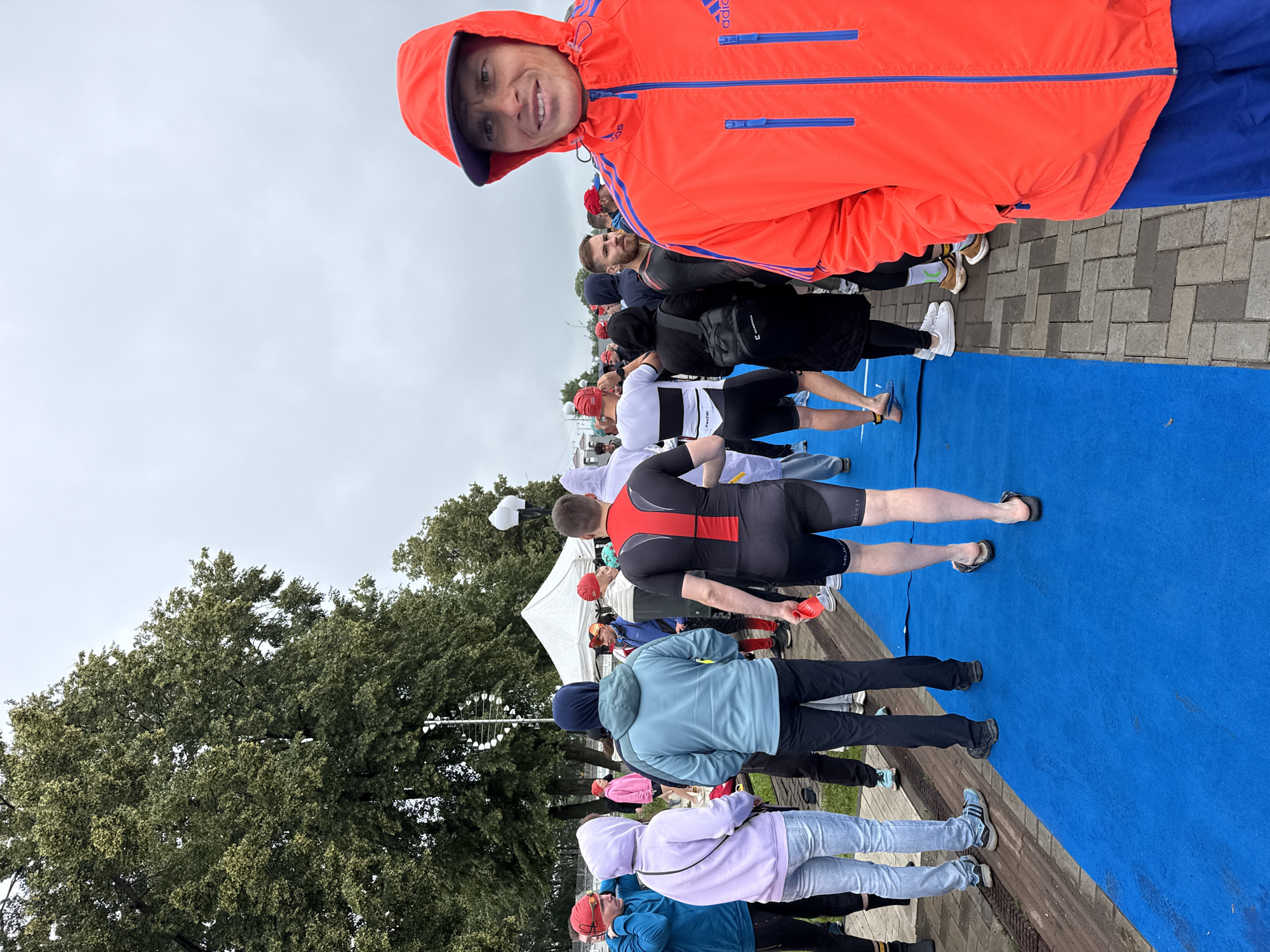 In Votkinsk, Russia, a crowd of people are gathered outdoors on a cloudy day, possibly for a sporting event or race. A vibrant blue carpet or mat runs across the foreground, with a brick-paved walkway bordering it. Many individuals are dressed in athletic wear, including wetsuits, t-shirts, and shorts. Some are wearing hats and some are not. In the foreground, a man in an orange hooded jacket with blue trim is prominently featured, looking towards the camera with a slight smile. Behind him, other participants are visible, some walking along the blue carpet, others standing and conversing. In the background, large trees with green foliage line the area, and a white tent is set up, suggesting a registration or staging area. The overall atmosphere appears to be one of anticipation and preparation.