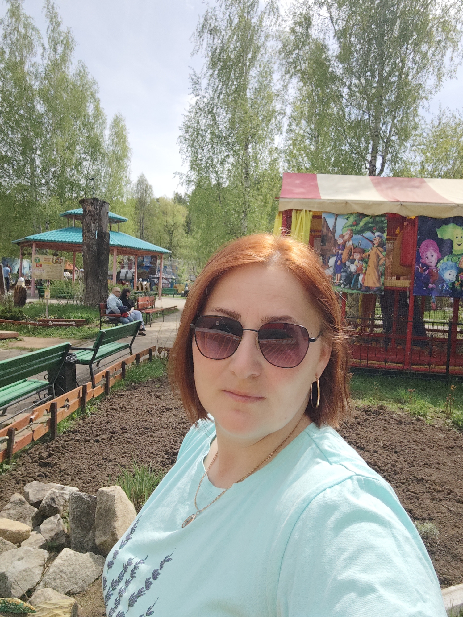 The image is a selfie taken outdoors in a park-like setting in the city of Chaikovsky, Russia. In the foreground is a woman with auburn hair, wearing sunglasses, a light blue t-shirt, and gold jewelry (earrings and a necklace). She is looking directly at the camera. Behind her, the ground is mostly bare soil with some rocks and grass. There are green benches and a low wooden fence.
In the midground, there is a blue-roofed gazebo, with people visible sitting on benches around it. There is also a small, colorful booth with cartoon images on its sides, suggesting it may be a children's attraction.
The background consists of trees with light green foliage, indicating it may be spring or early summer. The sky is overcast. The scene suggests a public park or recreational area.
