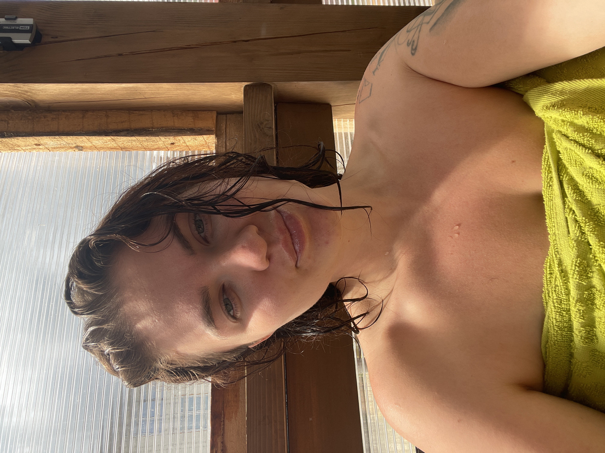 A person with wet hair and a tattoo on their left shoulder is partially visible in the image, wrapped in a green towel. The person is positioned in what appears to be a sauna or wooden structure, with light shining through the wall behind them. The location "Мосметаллоконструкция Онт, Russia" is not visible in the image.