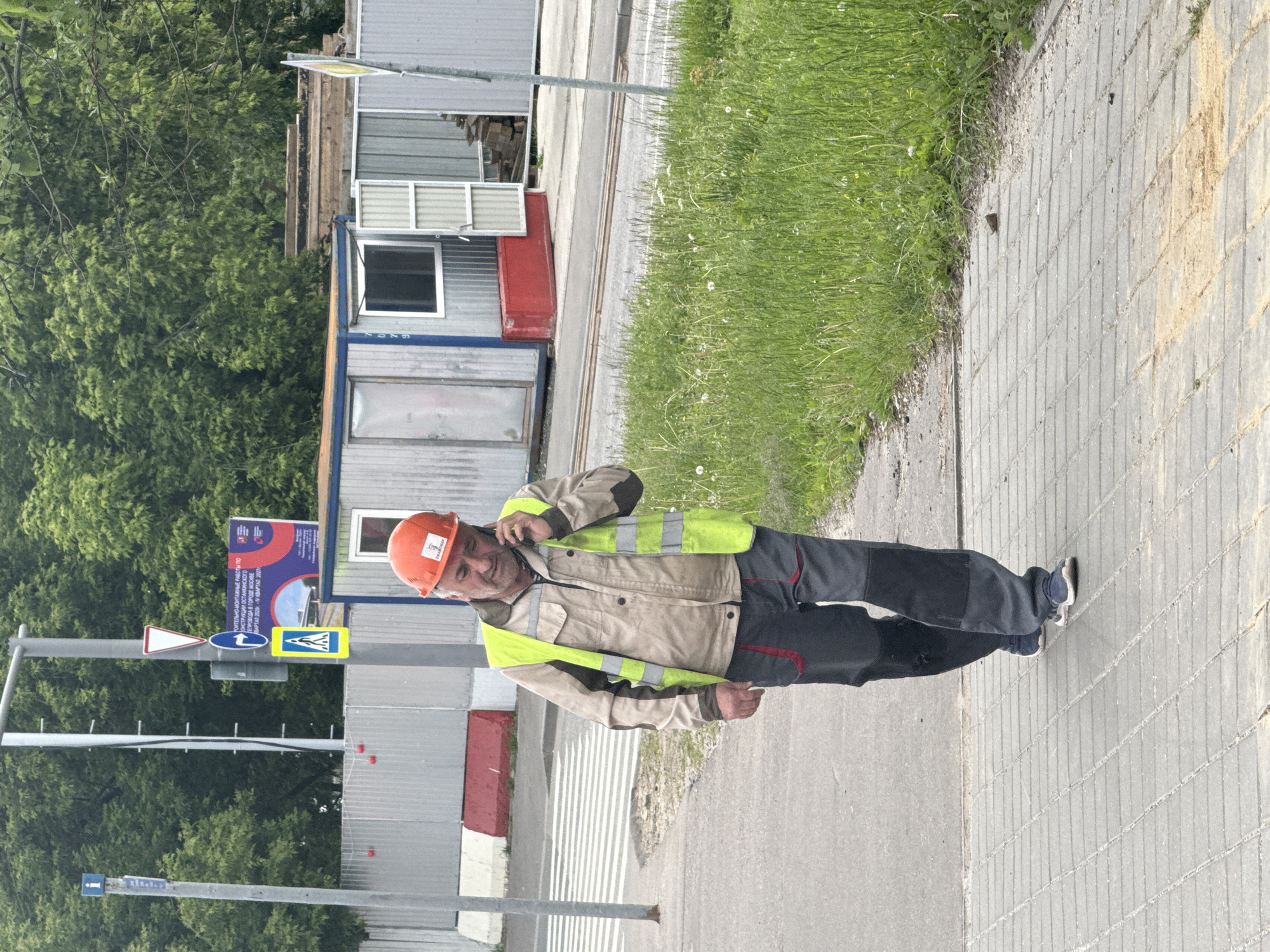 The image captures a daytime scene in Moscow, Russia, depicting a man engaged in a phone conversation near a construction site. The man, a worker, is standing on a paved sidewalk, wearing an orange hard hat, a beige work jacket, a yellow-green high-visibility vest, and dark work pants. He holds a phone to his right ear and looks towards the camera with a slight smile.
To his left, a lush green treeline is visible, while behind him are several temporary modular buildings, resembling construction site offices or storage containers, colored in grey, red, and blue. The ground beneath him transitions from the paved sidewalk to a strip of green grass and then an asphalt road.
A metal pole with multiple Russian road signs stands prominent on the left. These include a "give way" sign (inverted red triangle), a blue circular sign with a white arrow (likely indicating a mandatory direction or a general direction), a yellow square with a black pedestrian crossing symbol, and a blue rectangular information sign. A large blue informational sign is also visible in the background, partially obscured, which reads: "СТРОИТЕЛЬНО-МОНТАЖНЫЕ РАБОТЫ ПО КОНСТРУКЦИИ ОСТАНКИНСКОГО ТЕЛЕПРОВОДА В ГОРОДЕ МОСКВЕ С 4 КВАРТАЛА 2025 - 4 КВАРТАЛ 2027". This text translates to "Construction and installation works for the Ostankino TV Tower structure in the city of Moscow from Q4 2025 - Q4 2027," confirming the location and the nature of the activity in the vicinity. The lighting suggests an overcast day, providing soft, even illumination.