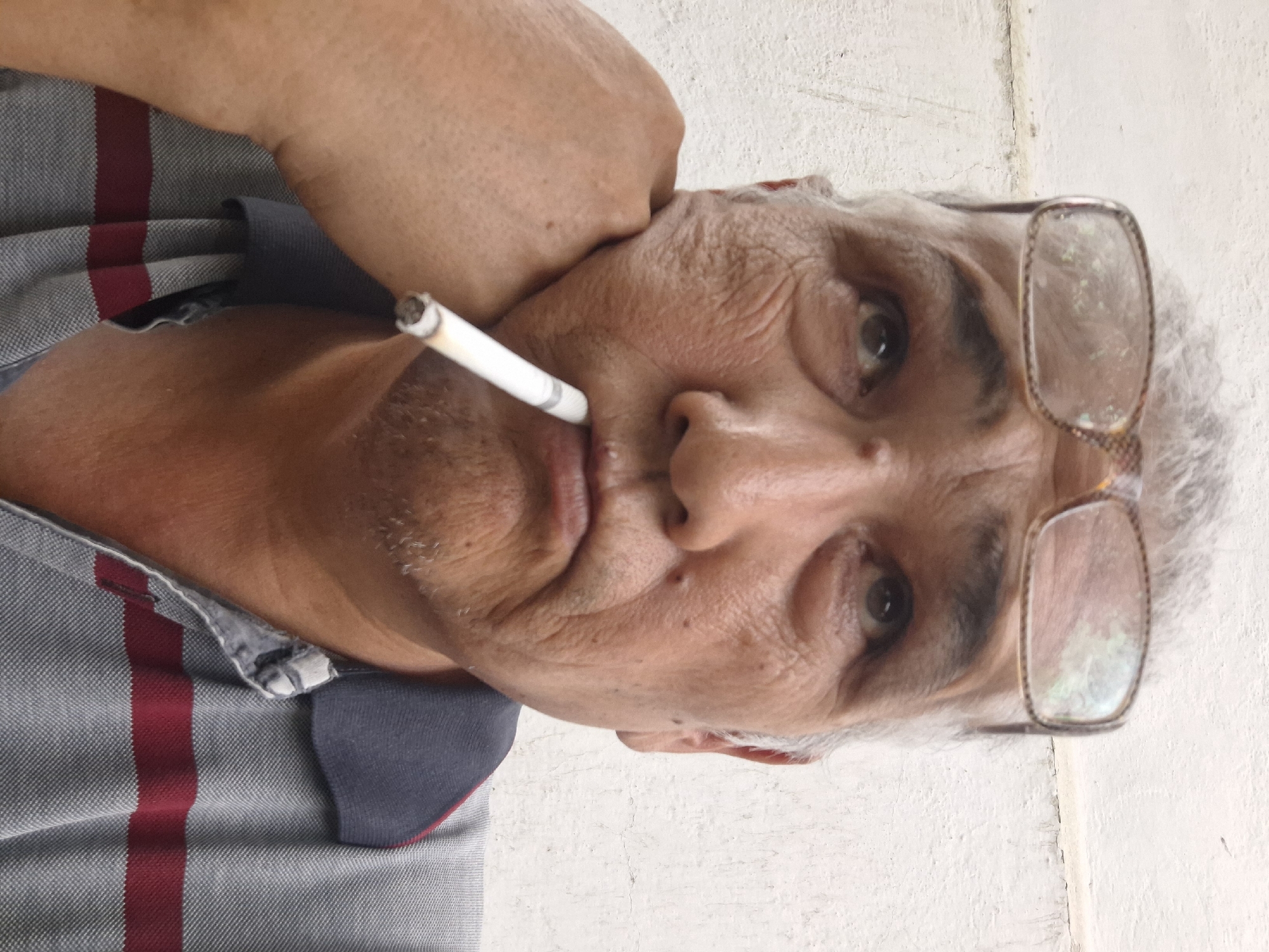A middle-aged man with graying hair and glasses rests his chin on his left hand. He has a cigarette hanging from his lips, angled upward. The man wears a gray polo shirt with two horizontal maroon stripes across the chest. The background is a plain, textured white wall, suggesting an indoor or sheltered outdoor setting. There is no specific indication of the city of Almaty, Kazakhstan, in the image.