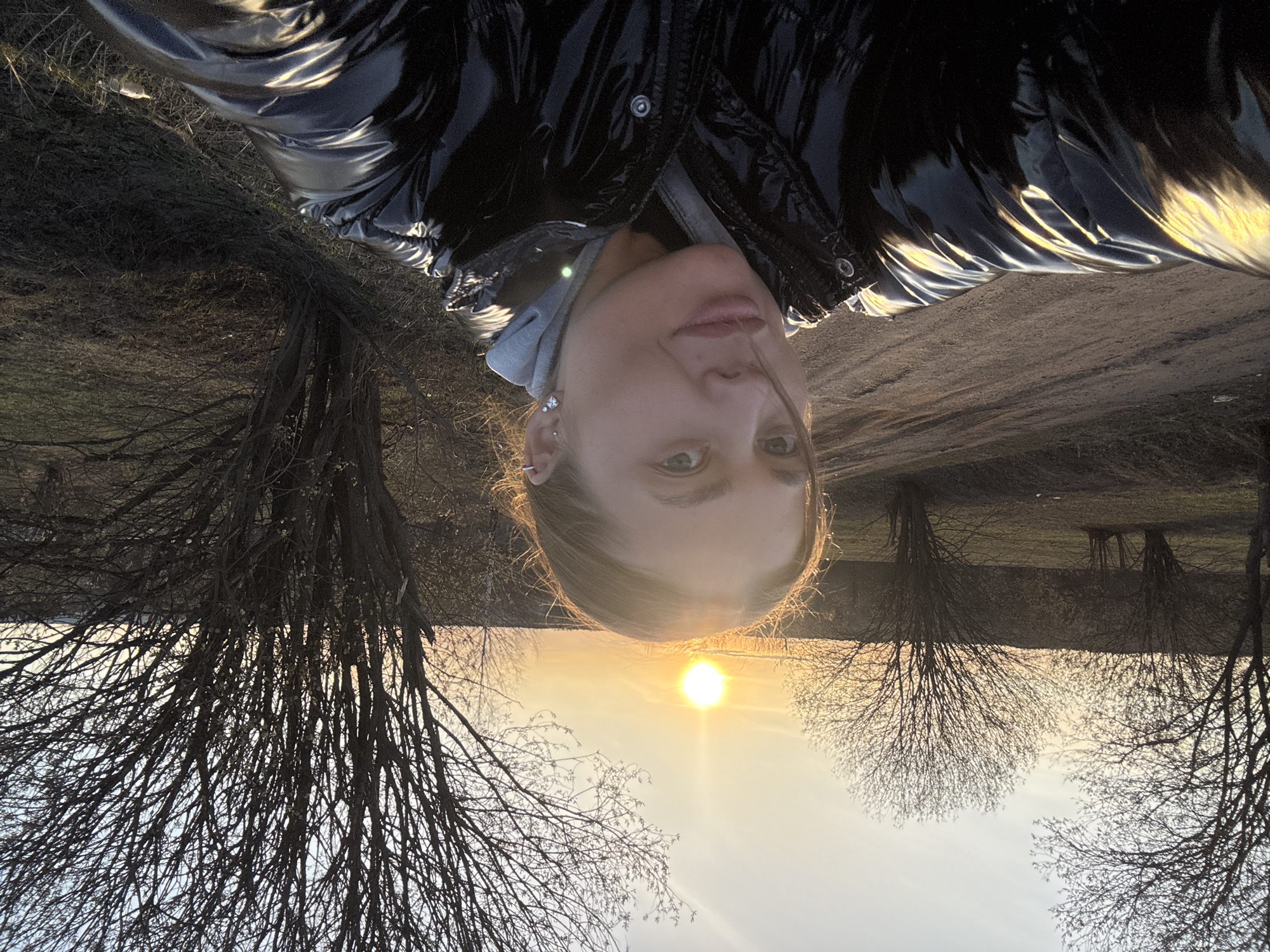 A selfie portrait is taken outdoors, with the camera rotated 180 degrees. The subject is a woman with fair skin and light brown hair, wearing a black, shiny puffer jacket with a grey hooded sweatshirt underneath. The background features a landscape with bare trees silhouetted against a bright, golden sky where the sun is setting. The terrain appears to be fields or open land. The location is not identifiable.