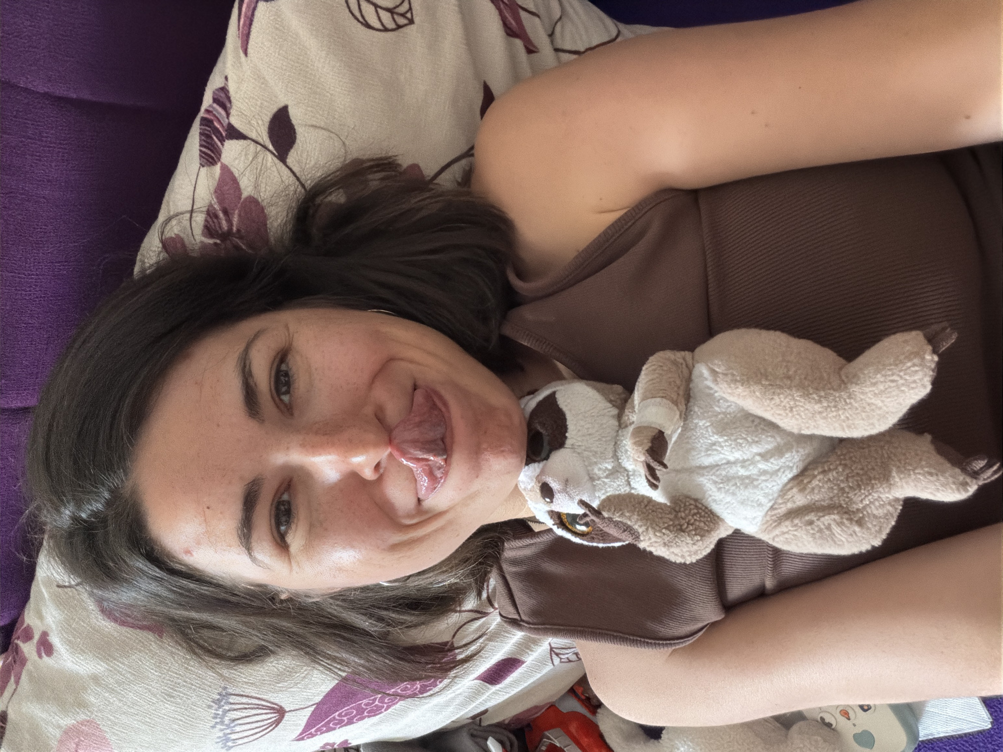 A young woman with dark hair is lying on her back with her tongue sticking out. She's wearing a brown tank top and lying on a pillow with a floral pattern. A stuffed sloth is resting on her torso. The background includes a purple couch. There is no visual information to determine if this image was taken in Budva, Montenegro.