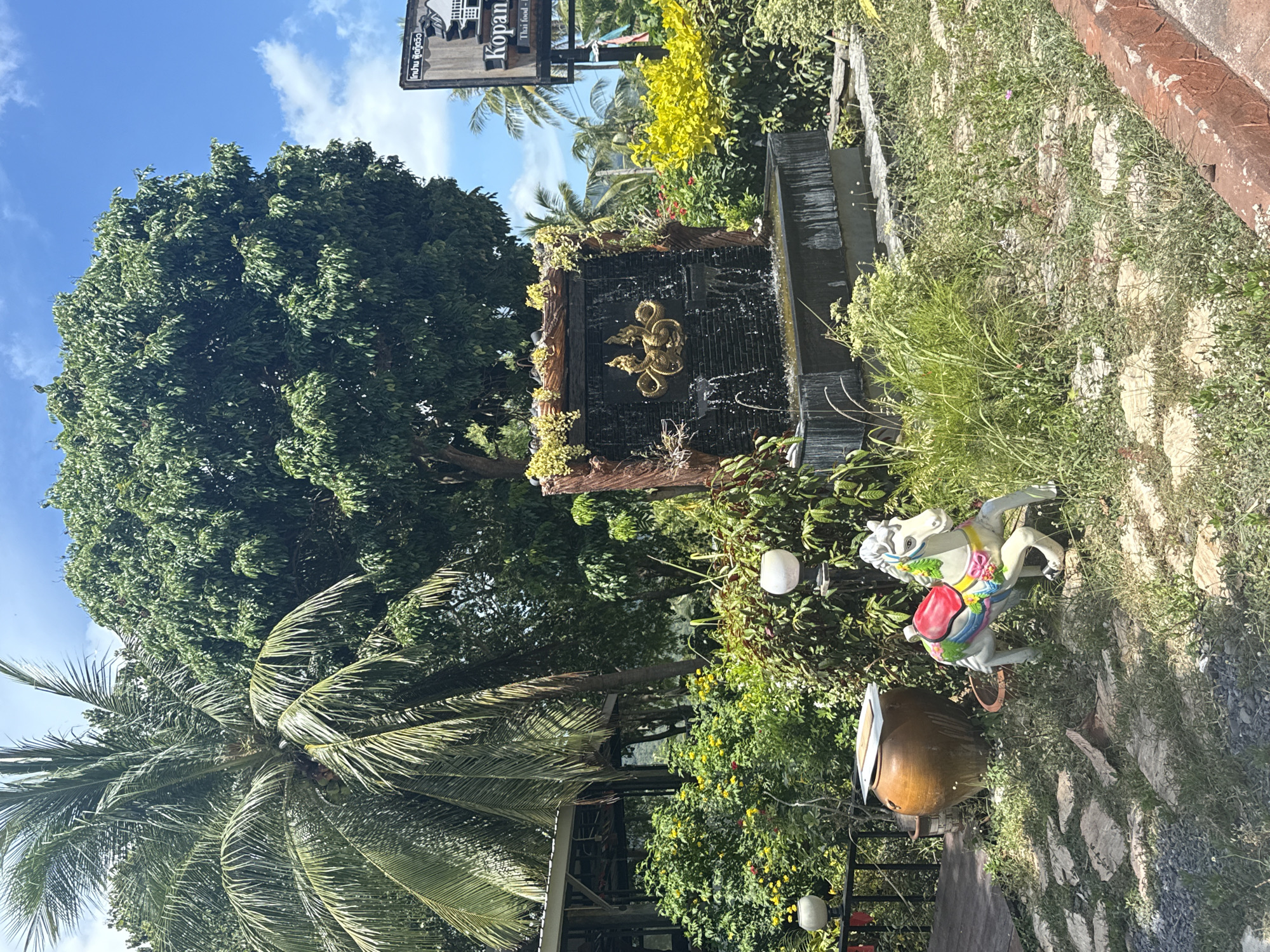 An outdoor scene in Ban Tai Subdistrict, Thailand, features a decorative fountain with golden serpents as its centerpiece, surrounded by lush greenery and tropical plants. In the foreground, a colorful, ceramic horse statue stands on a path of natural stones. A large, brown ceramic pot is visible to the left of the horse. In the background, a tall palm tree and a dense canopy of green leaves dominate the view, with a glimpse of a building structure to the left. A sign for "Kopan Thai Food" is also visible on the right side of the image. The sky is clear blue with scattered white clouds.