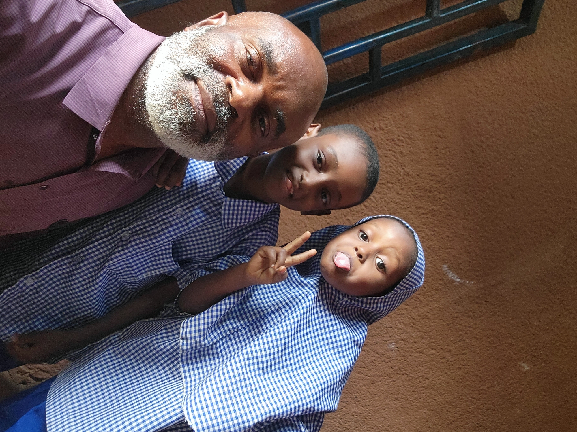 A selfie shot from above captures a man and two children in Maiburiji, Nigeria. The man, light-skinned with a gray beard and shaved head, wears a burgundy checkered shirt. The two children, dressed in blue and white checkered school uniforms, are positioned below him. One child makes a peace sign and sticks out their tongue. The background is a brown wall with a dark blue railing visible near the top.