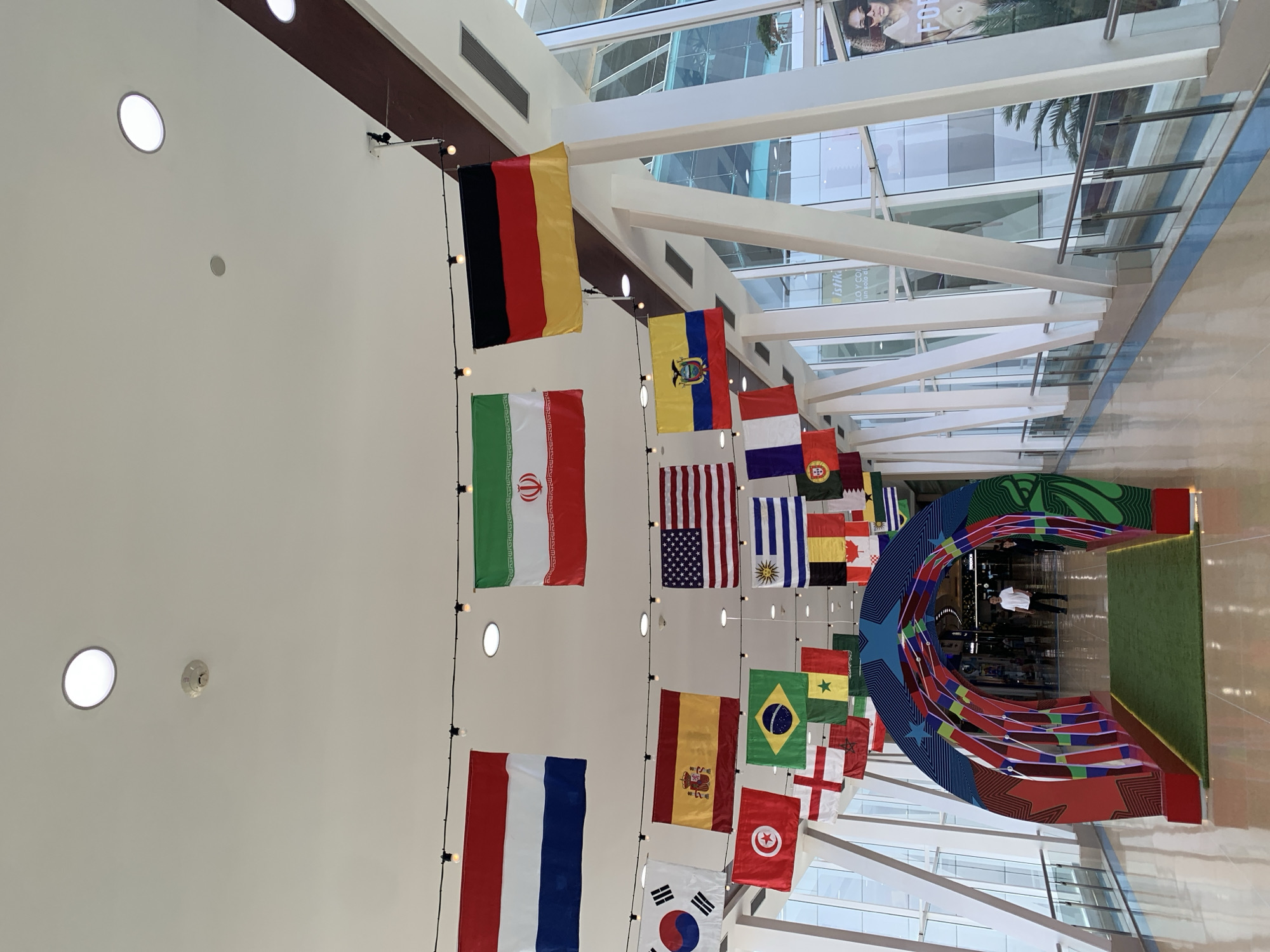 The image depicts an indoor walkway or mall with numerous national flags suspended from the ceiling by black wires. The flags are arranged in rows and appear to represent various countries. A brightly colored, arch-shaped entrance decorated with geometric patterns and stars is positioned in the foreground. A green carpeted path leads into the archway. Two individuals are visible in the distance, walking through the arch. The walkway is characterized by white structural beams, large glass windows revealing an outdoor environment with vegetation, and reflective flooring. The ceiling features recessed lighting and what appears to be a smoke detector. Based on available information, the scene is located in Porlamar, Venezuela, specifically at the Sambil Margarita mall, which is known for displaying flags of different nations as decorative elements.