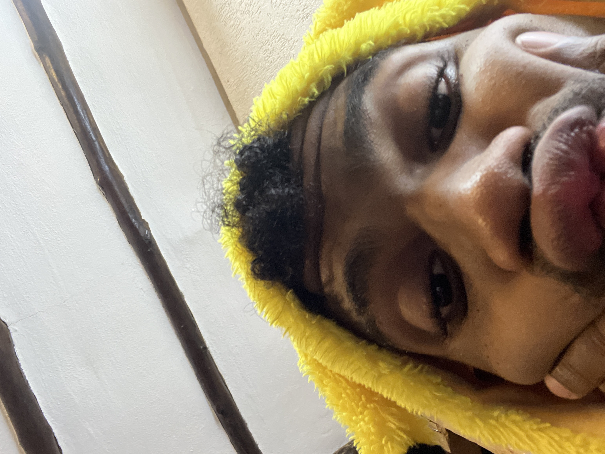 A close-up, angled shot of a person with dark skin and short, curly black hair. They are wearing a yellow hooded garment. Their lips are pursed, and one hand is touching their face. The background shows a textured white wall with dark vertical stripes.
The image lacks sufficient detail to determine the location in Porlamar, Venezuela.