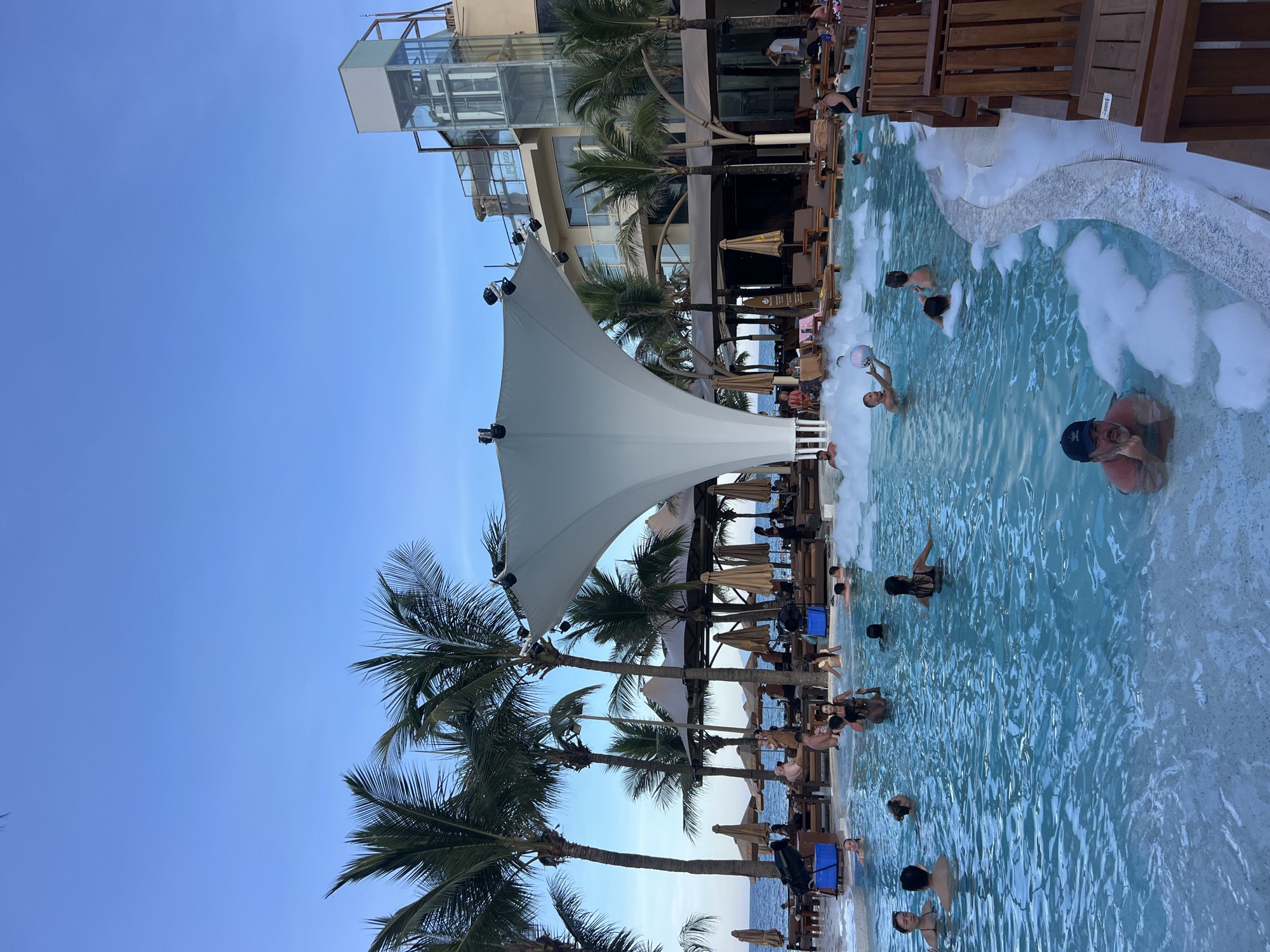 An outdoor swimming pool area at a resort is depicted under a clear blue sky. Palm trees line the background, with a modern building visible behind them. In the pool, several people are enjoying the water, some playing with a beach ball and others relaxing in the foam that covers parts of the pool. Lounge chairs and umbrellas are set up around the pool. The scene appears to be a lively and relaxing vacation spot.