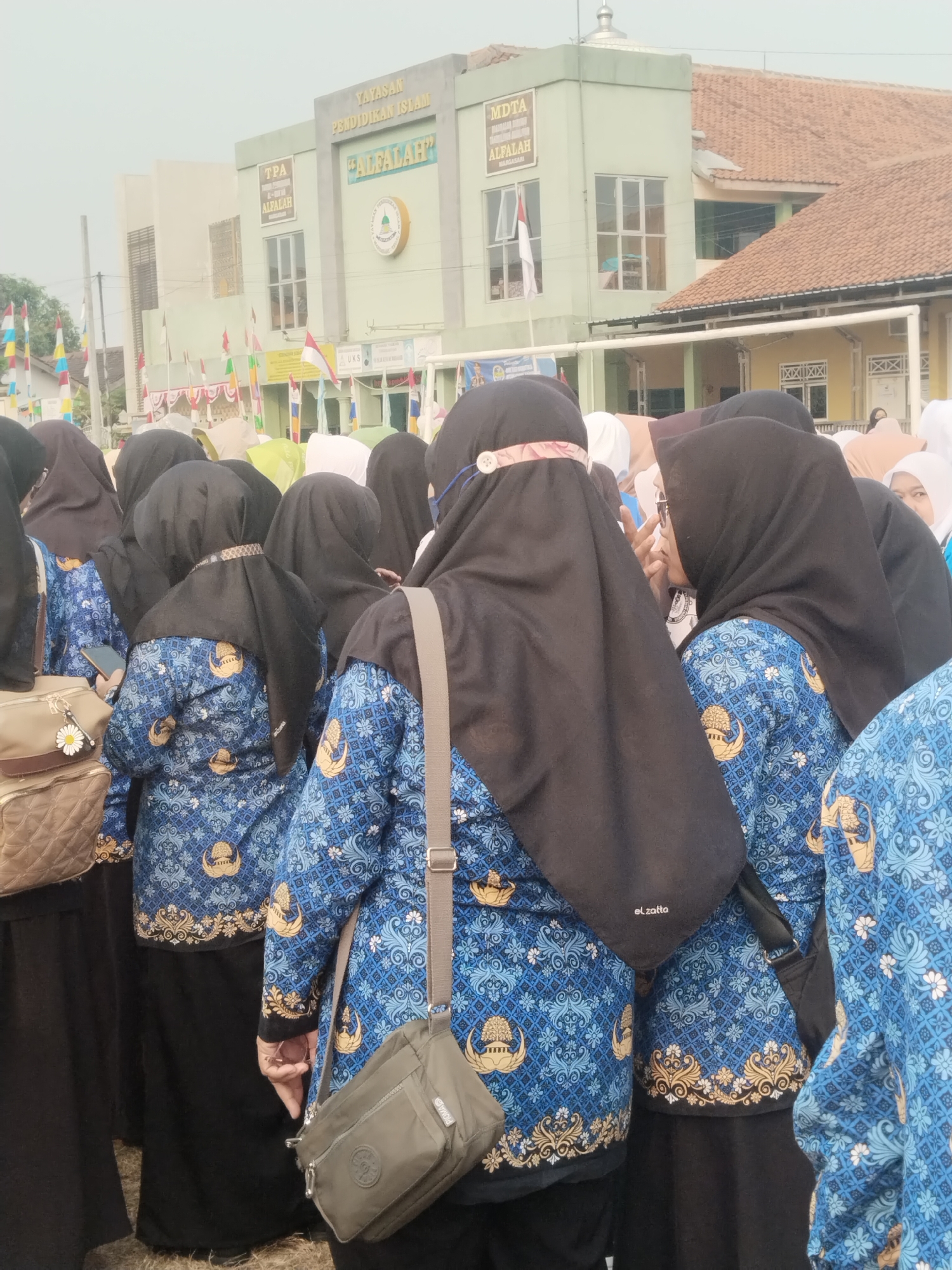 The image shows a group of women, likely teachers or staff, gathered outside a building that appears to be a school or educational institution in Margasari, Indonesia. The building has a light green facade with signage indicating "YAYASAN PENDIDIKAN ISLAM" and "AL FALAH." Several women are wearing matching blue uniforms with a batik-like pattern, featuring national symbols and gold embellishments. They are also wearing hijabs, predominantly black. Some women are holding bags. The background includes other buildings, a flagpole with Indonesian flags, and a cloudy sky. The overall scene suggests an outdoor event or gathering related to the educational facility.