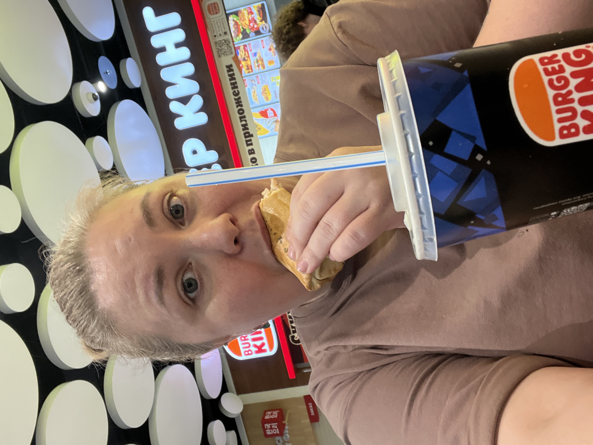 A woman is eating a sandwich and holding a Burger King drink at a Burger King restaurant in Sochi, Russia. She is wearing a brown shirt. The restaurant has white circular decorations on a black wall. A menu board is visible in the background.