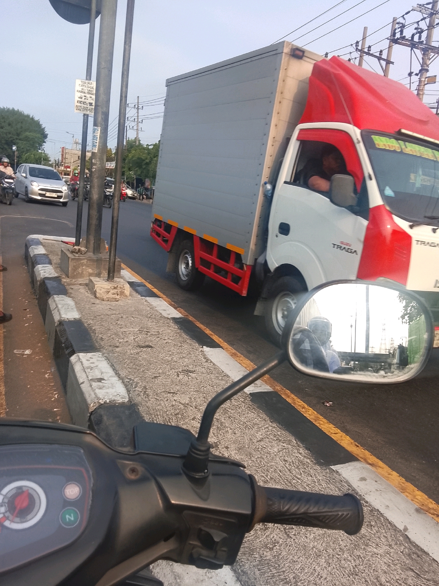 This is a ground-level, first-person perspective shot, taken from a motorcycle, looking forward and slightly to the right. The shot is composed of a busy street scene in Medaeng, Indonesia. In the foreground, the handlebars and part of the dashboard of the motorcycle are visible. The dashboard displays a speedometer with a red needle, and indicator lights labeled "TOP" and "N".
Dominating the mid-ground is a large, white and red box truck, with "ISUZU TRAGA" branding visible on its side. The truck is in motion, as indicated by its blurred appearance and the open driver's side window. A person, likely the driver, is partially visible inside the truck cabin. The truck is on the right side of the road.
To the left of the truck, a silver hatchback car is also in motion. Several other motorcycles and a few pedestrians are present on the road and sidewalk, suggesting moderate traffic. The road is paved, with yellow and white lines marking the lanes and edge. A raised concrete curb with black and white markings runs along the side of the road, separating it from a pedestrian walkway.
In the background, utility poles with power lines are visible against a bright sky. Trees line the far side of the road. A sign is mounted on a pole near the left edge of the frame, displaying text in Indonesian. The overall impression is that of a typical urban street scene in Indonesia during daylight hours.