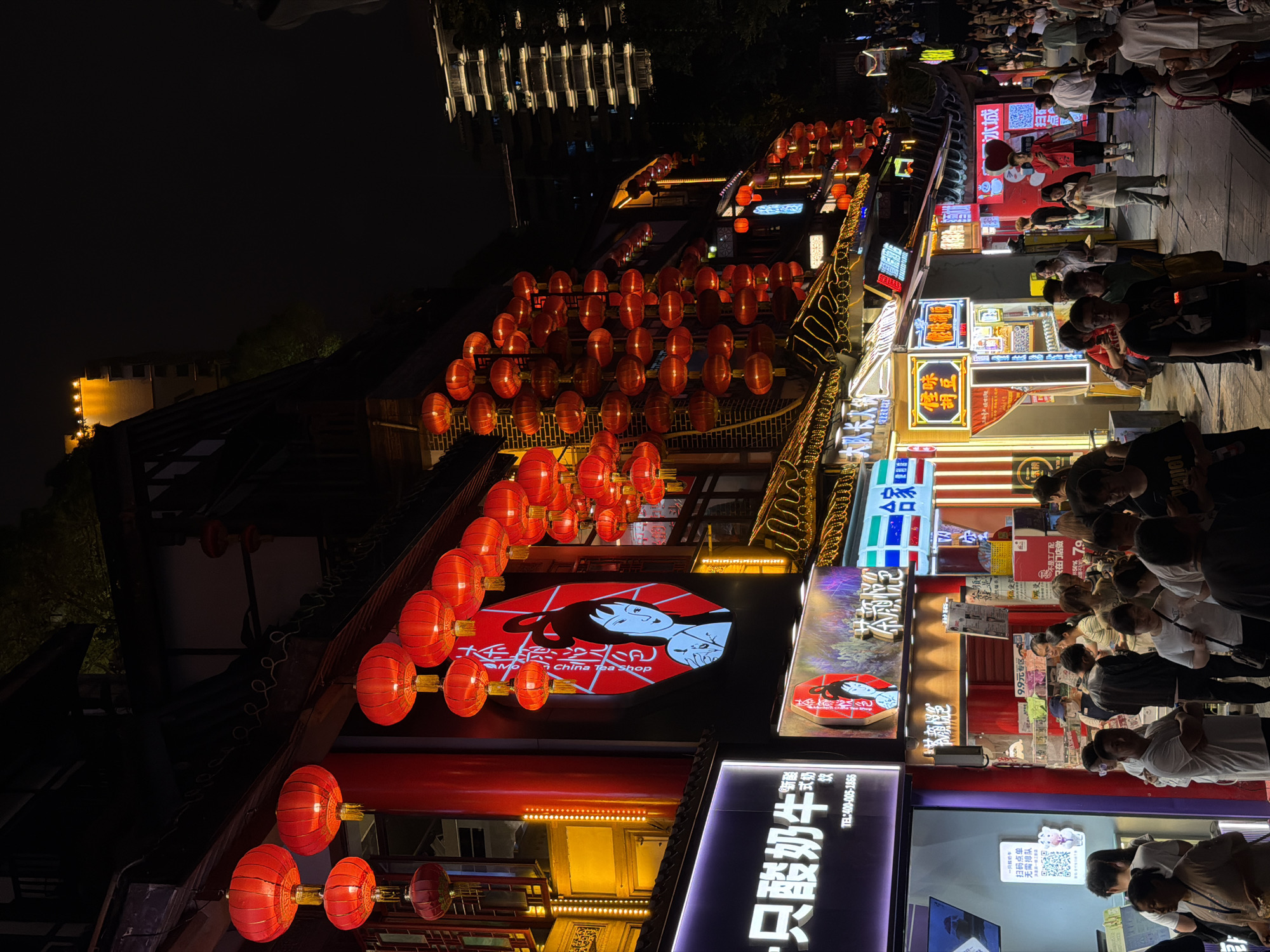 This is a nighttime scene in Yuzhong District, China, depicting a bustling street or market area illuminated by numerous red lanterns and neon signs. The image is oriented vertically, with the bottom of the scene at the top of the frame. A crowd of people is visible, browsing shops and moving along a paved walkway. The shops are adorned with various signs, some in Chinese characters and others with English text, indicating businesses like "China Tea Shop" and "大队长火锅" (Captain Hot Pot).
The atmosphere appears lively and vibrant, typical of a busy evening in an urban commercial district. The red lanterns create a warm and festive ambiance, contrasting with the dark night sky. Several shops display promotional posters and screens, with one sign advertising "9.9元专区" (9.9 Yuan Special Zone). The people are dressed in casual attire, suggesting a relaxed shopping or dining experience. The overall impression is one of a dynamic and engaging street scene, filled with activity and commercial life.