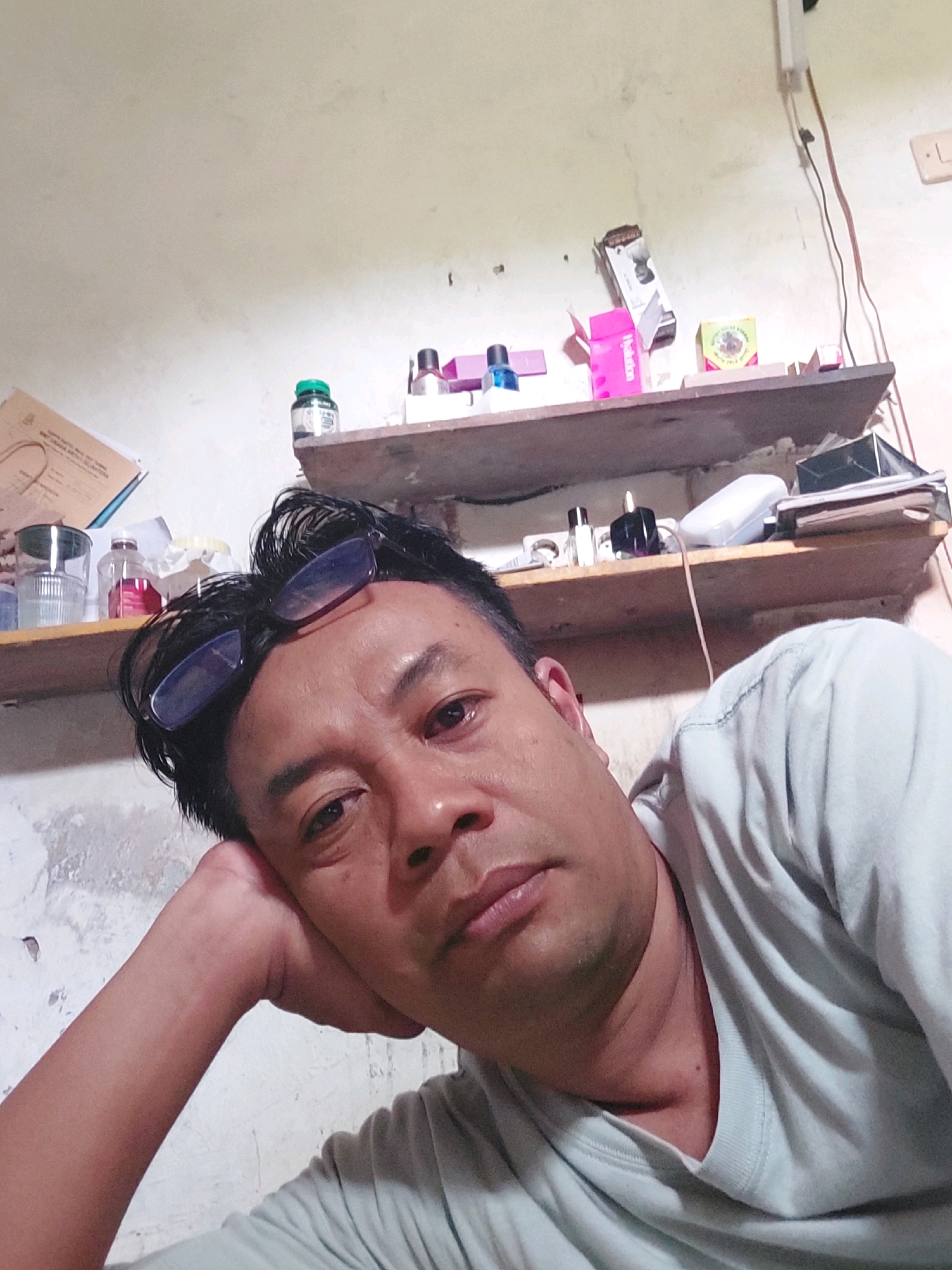 A medium shot captures a man resting his head on his arm, looking towards the camera. He has dark hair and is wearing a light-colored t-shirt. His glasses are perched on top of his head. In the background, two shelves are visible, cluttered with various items including bottles, boxes, and papers. The walls appear to be textured and somewhat aged. The image is likely taken indoors, possibly in a residential setting in Sarangan, Indonesia, given the context provided.