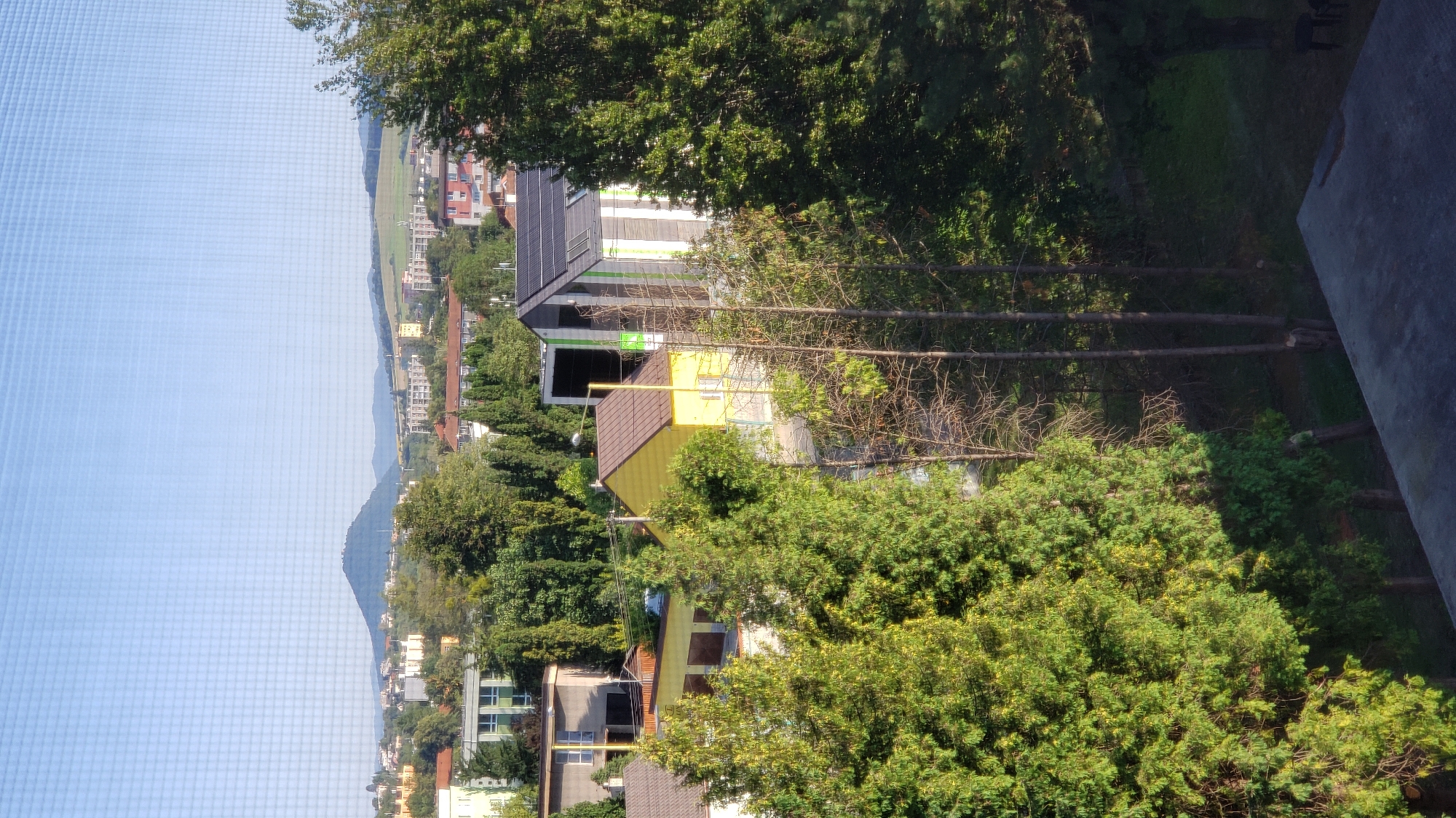 The image captures a view of the city of Prešov, Slovakia, looking towards the Slanské vrchy mountains. In the foreground, lush green trees dominate the scene, with a concrete structure, possibly a balcony or roof, visible in the bottom right corner. A bright yellow house with a brown roof stands out amidst the greenery. To its right, a modern building with a gray and white striped facade and solar panels on the roof is partially visible. In the middle ground, several residential buildings with red roofs and apartment blocks can be seen, interspersed with more trees. In the background, a prominent, rounded mountain, likely one of the Slanské vrchy, rises against a clear blue sky. The overall impression is of a suburban or residential area within the city, with a natural landscape on the horizon. There are no people or animals visible in the image.