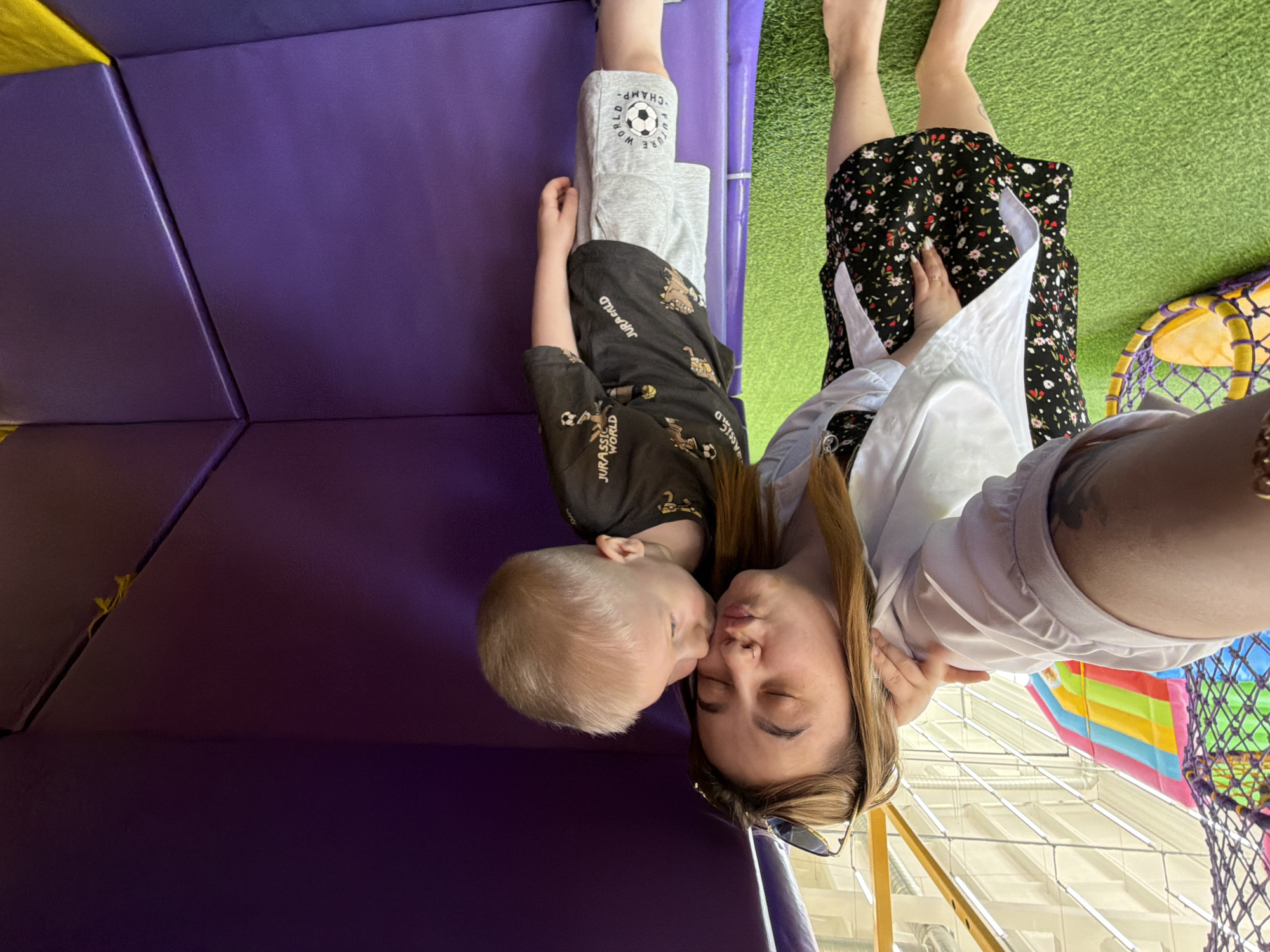 A woman and a young child are positioned head-to-head in what appears to be an indoor play area. The woman has her eyes closed and is wearing a black floral dress with a white shirt over it. The child, wearing a "Jurassic World" t-shirt and gray pants with a soccer ball design, is kissing the woman's cheek. They are surrounded by play area surfaces with vibrant colors: purple, green, and yellow. A play structure with netting is visible. The location is Chaikovsky, Russia.