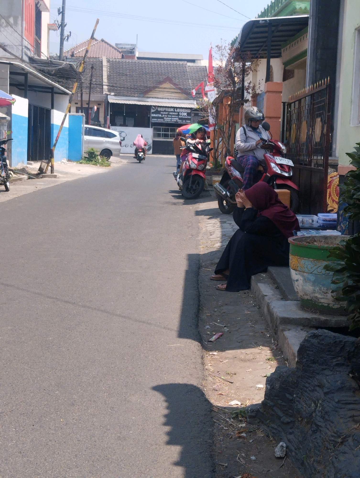 In Malang, Indonesia, on a sunny day, a street scene unfolds. A woman in a maroon hijab and black abaya is seated on the curb, her hands clasped over her face, suggesting distress. She is near a red motorcycle parked beside a dark grey, textured object that appears to be a decorative rock or sculpture. A person in a helmet, riding a red scooter, is visible behind her, looking down at their phone. Another red motorcycle is parked further up the street, with two people on it, one wearing a pink hijab. A white car is parked on the opposite side of the road. The street is paved, and there's a sidewalk with a drainage ditch. Buildings line the street, featuring various architectural styles, including one with a prominent red and white striped pole. The overall impression is a snapshot of daily life in a bustling Indonesian city, with a hint of individual emotion captured amidst the activity.