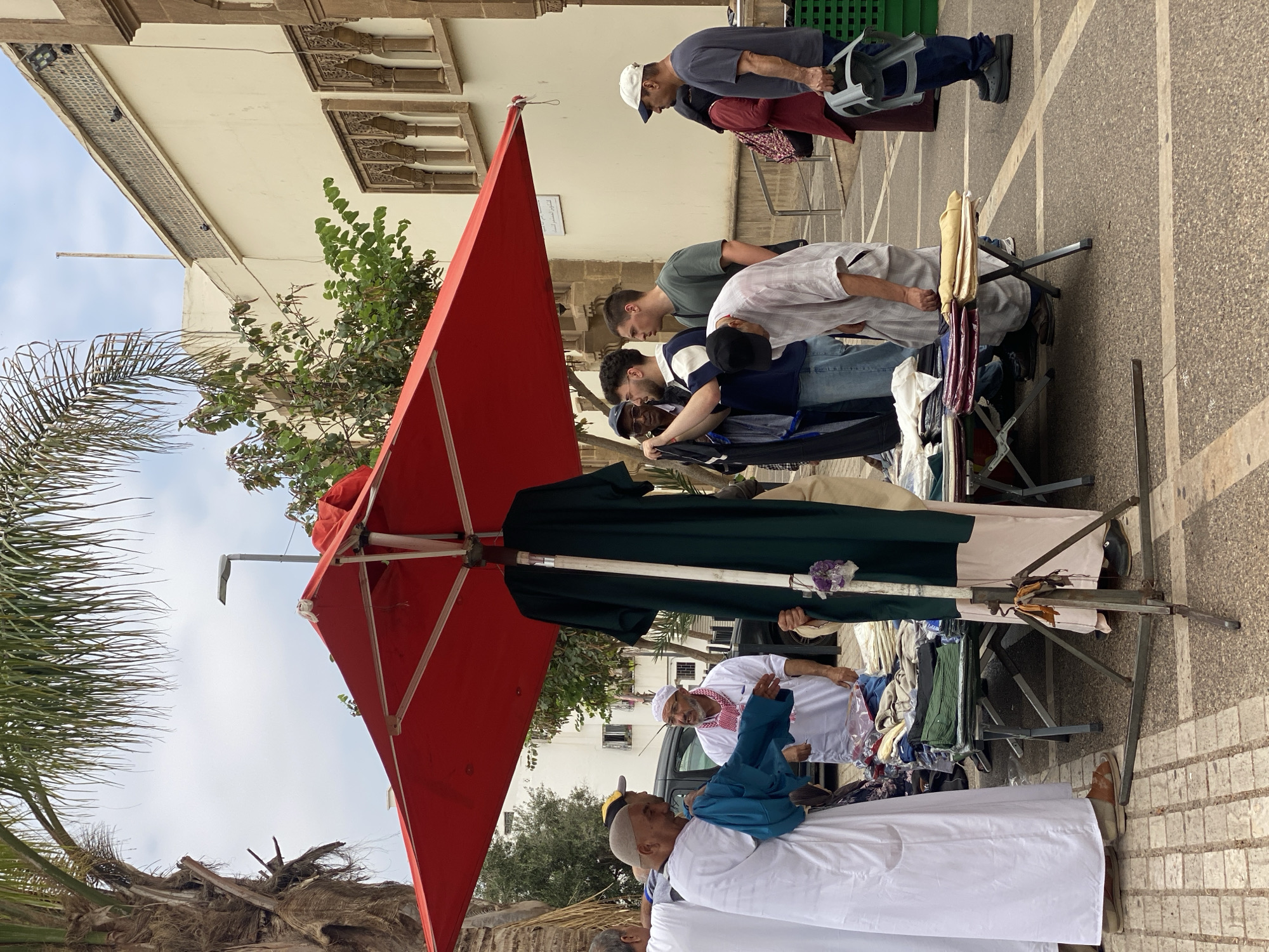 This image depicts a bustling outdoor market scene in Agadir, Morocco, likely during the daytime under an overcast sky. The perspective is somewhat immersive, placing the viewer among the shoppers and stalls.
In the foreground and midground, several men are gathered around multiple market stalls. These stalls, covered with various garments and fabrics for sale, are shaded by a large, bright red umbrella that dominates the left side of the frame. Shoppers, dressed in a mix of traditional attire such as white thobes and keffiyehs, and more casual clothing like t-shirts and jeans, are seen browsing and interacting with the merchandise. One man in a white thobe and blue top has his back partially to the camera, while another in a white thobe and a red and white checkered keffiyeh smiles towards the viewer.
The ground is paved with patterned tiles or cobblestones, featuring distinct linear designs. In the background, multi-story buildings with traditional Moroccan architectural elements are visible, characterized by ornate carvings around windows. Green trees and the fronds of a palm tree provide natural elements to the urban setting. The overall atmosphere suggests a typical, lively local market where people are engaged in shopping and commerce. No legible text is visible in the image.