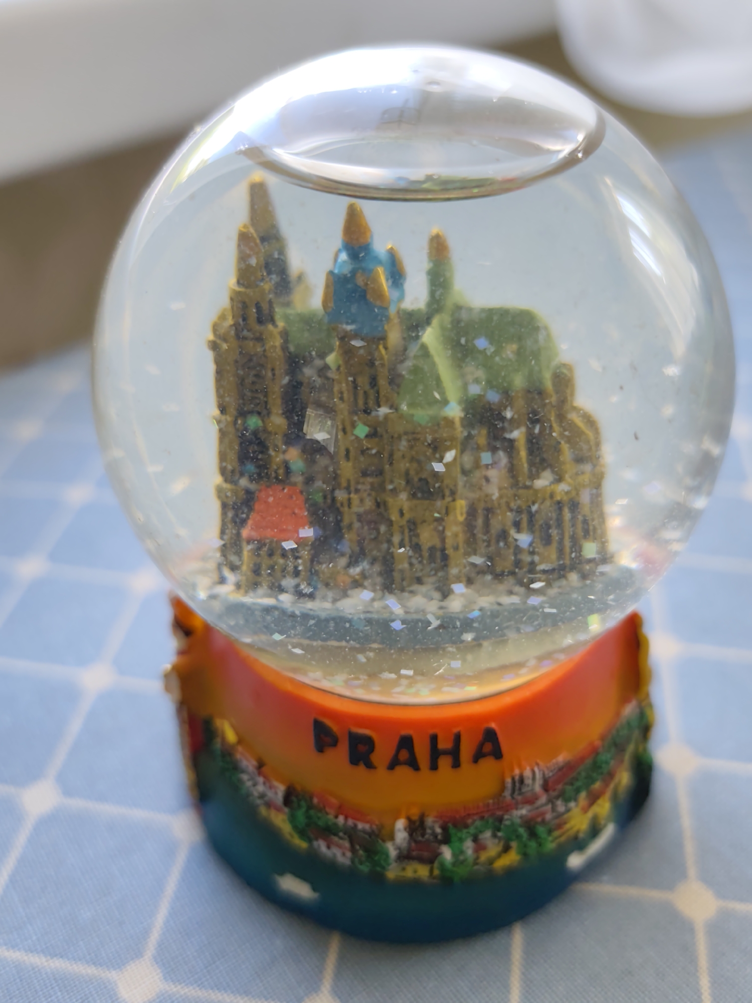 The image displays a snow globe with a miniature cityscape inside. The cityscape appears to be a historical or architectural site with tall, narrow buildings and a blue dome. The snow globe itself is filled with clear liquid and contains scattered glitter. The base of the snow globe is orange and teal, decorated with an image of a city skyline and the word "PRAHA" in black lettering. The snow globe rests on a light blue surface with a white grid pattern. There is no indication of the location being in Бараўлянскі сельскі Савет, Belarus.