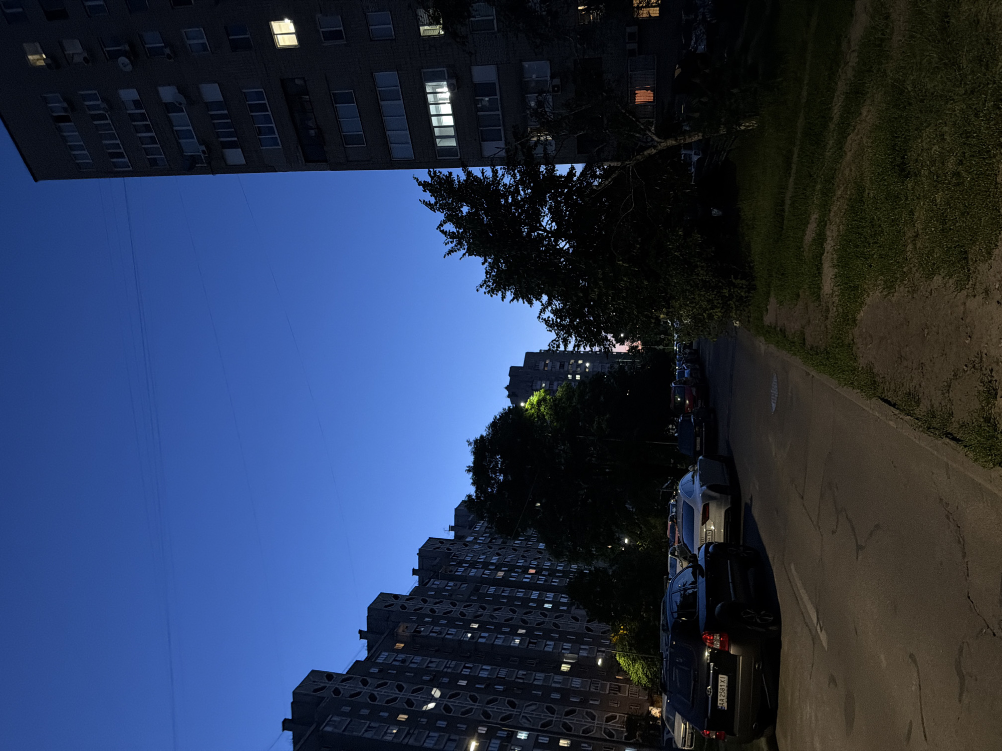 The image captures a residential street scene in Kyiv, Ukraine, during dusk. A row of apartment buildings, characteristic of Soviet-era architecture with distinctive circular patterns on their facades, lines the left side of the street. The buildings are illuminated by lights from some of the windows, suggesting occupancy. On the right, a taller, more modern apartment building stands, also with illuminated windows. A line of parked cars is visible along the street, including a black SUV in the foreground with a Ukrainian license plate. Trees and grassy areas are interspersed between the buildings and the street, adding greenery to the urban landscape. The sky is a deep blue, indicating the transition from daylight to night, with a few faint horizontal lines suggesting power or telephone wires. The overall impression is a quiet evening in a typical Kyiv residential district.
