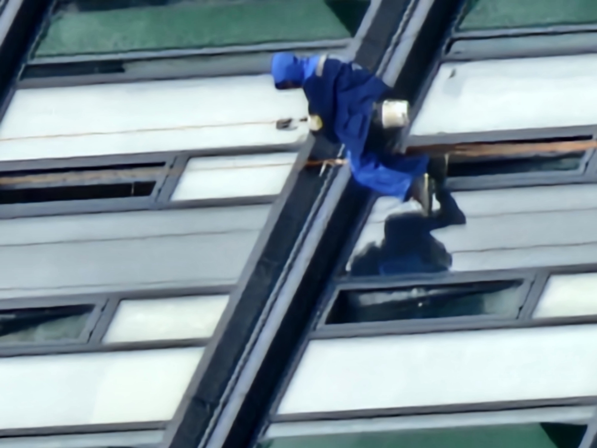 This media file captures a high-altitude window cleaner working on the exterior of a modern building in Moscow, Russia. The subject is a lone individual wearing a full blue uniform, including a hood, and is suspended by ropes and harnesses, demonstrating a potentially hazardous work environment.
The scene is characterized by the repeating pattern of horizontal window panes, indicative of a large commercial or residential building. The glass surfaces reflect the sky, suggesting it is daytime. The angle of the shot, looking up at the worker, emphasizes the height and the scale of the building. The lighting suggests it could be a bright day, with some glare on the windows.
The activity is clearly a window cleaning operation. The subject is positioned on a rope system, equipped with tools and cleaning supplies, though these are not highly detailed due to the image quality and distance. The worker's posture suggests focus and concentration on the task at hand.
Notable details include the uniform, which appears to be standard workwear for such professions. The building's architecture seems contemporary, with dark framing around the windows. A shadow of the worker is visible on the building's facade, further reinforcing the daytime setting and the sun's position. No specific text is discernible in the image. The overall impression is one of a challenging but routine task being performed in an urban environment.