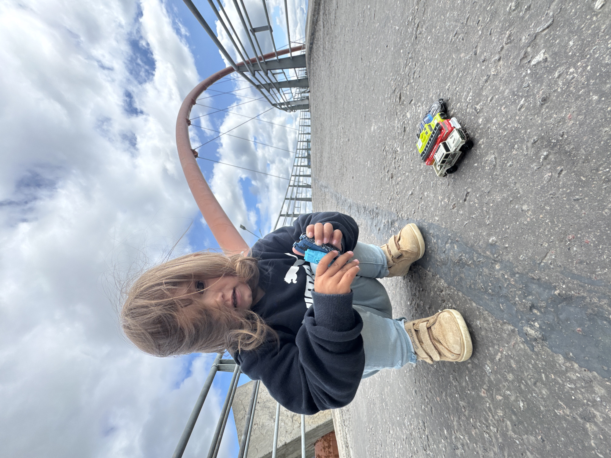 An undated, eye-level, full-shot photograph captures a young child, with windswept blonde hair obscuring part of their face, crouching on the asphalt surface of a pedestrian bridge. The child, wearing a dark blue Puma hoodie, light blue jeans, and tan Velcro sneakers, holds two small blue toy cars in their right hand. In front of the child, a row of three toy vehicles—a white pickup truck, a red fire truck, and a blue police car—are lined up on the bridge's surface. The bridge features a curved, coral-colored archway with suspended cables and metal railings. The background reveals a cloudy sky with patches of blue and glimpses of urban structures, suggesting a location within the city of Saint Petersburg, Russia.