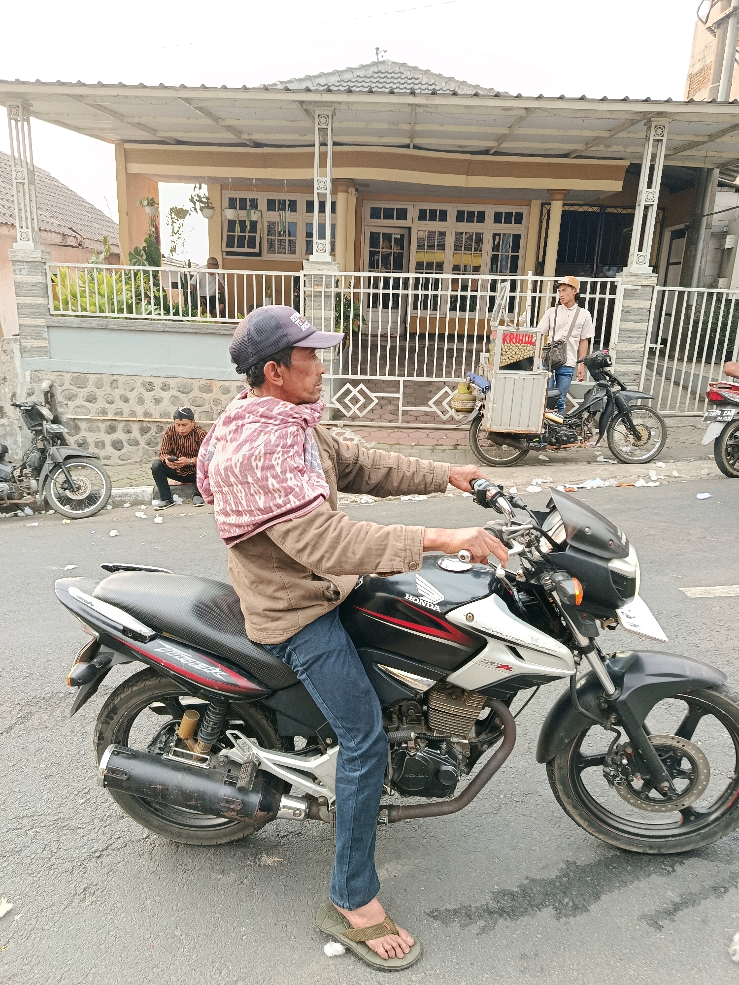 A man wearing a dark baseball cap, a light brown jacket, and blue jeans rides a black and silver Honda motorcycle. He has a pink and white patterned cloth draped around his shoulders. The motorcycle has a black exhaust pipe and a red stripe on the side. The man is looking to his right, away from the camera. In the background, a house with a white fence and a tiled roof is visible. Another man in a striped shirt and black pants is sitting on the ground in front of the fence, looking at his phone. A parked motorcycle is to the left of the man on the ground. The scene appears to be a street in Gubukklakah, Indonesia, based on the presence of a food cart with the word "KRIHUL" visible on it, which is a common Indonesian snack.