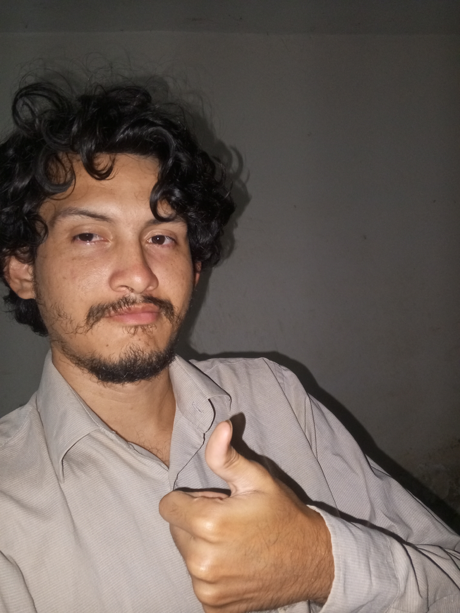 A man with dark, curly hair and a mustache gives a thumbs-up. He is wearing a light-colored collared shirt. The background is a plain gray wall. The location, Juan Griego, Venezuela, is not identifiable from the image.