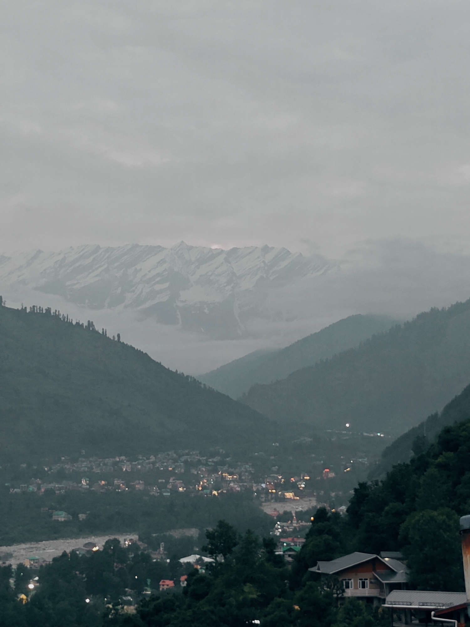 This media file depicts a panoramic view of a mountainous landscape under a cloudy sky. In the foreground, a cluster of houses and dense trees are visible, suggesting a settlement nestled within the mountains. The houses are partially illuminated, indicating it is either dusk or dawn.
The midground showcases rolling hills covered in vegetation, leading to a valley where a town or village is situated. The lights of the settlement twinkle through the trees, painting a picture of a warm community amidst the cool, subdued natural environment. A river or stream appears to flow through the valley, adding to the scenic beauty.
The background is dominated by majestic snow-capped mountains, their peaks partially obscured by clouds and mist. The sheer scale of the mountains evokes a sense of awe and grandeur. The overall atmosphere is serene and tranquil, with the overcast sky casting a soft, diffused light over the entire scene. The location is identified as Vashisht, India, a place known for its natural beauty and spiritual significance. The time of day appears to be either early morning or late evening, given the subdued lighting and the illuminated houses. The weather is overcast, contributing to the muted color palette of the image.