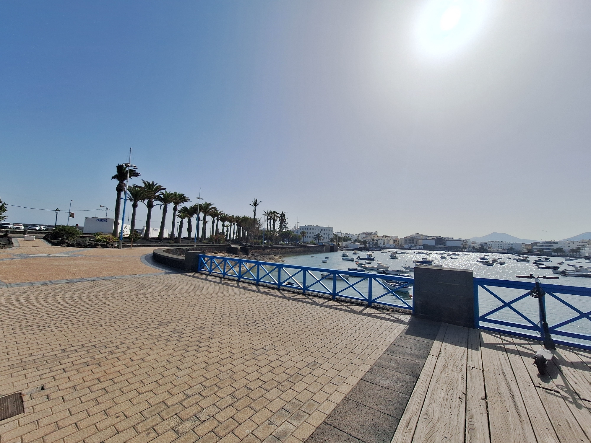 The image depicts a sun-drenched promenade in Arrecife, Spain, likely near the marina. In the foreground, a brick-patterned walkway stretches towards the right, transitioning to a wooden deck with an electric scooter parked on it. A vibrant blue railing with a criss-cross design borders the walkway, separating it from the calm body of water.
The water is dotted with numerous small boats, many of them sailboats, anchored in a harbor. Beyond the water, a townscape unfolds with low-rise buildings and some taller structures, including what appears to be a hotel or apartment complex. Palm trees line the promenade on the left, adding to the tropical atmosphere. The sky is a clear, bright blue, with the sun prominently positioned in the upper right quadrant, casting a strong glare. The overall scene suggests a pleasant day in a coastal city, perfect for leisure activities.