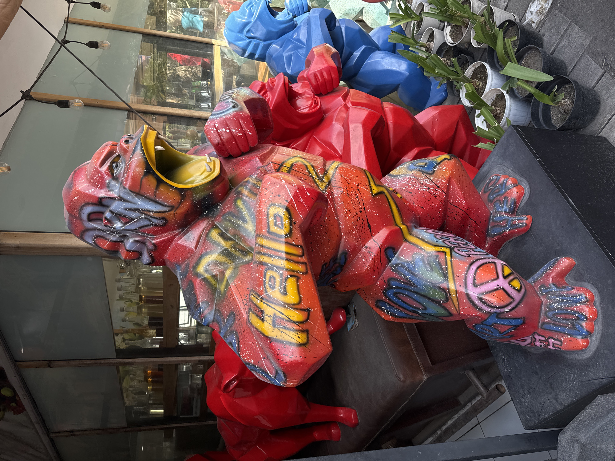 This image shows a vibrant street art display featuring large, polygonal gorilla sculptures in the city of North Kuta, Indonesia. The central sculpture is a red gorilla adorned with graffiti that spells out "HELLO" in yellow and features a peace sign on its leg. To the right, a bright blue gorilla sculpture is visible, also with its mouth open. Further to the left, a red, unpainted gorilla sculpture is partially visible. These sculptures are placed outdoors, possibly on a sidewalk or patio area, with potted plants arranged in front of them. Behind the sculptures, a building with large glass windows reveals shelves stocked with various bottles, suggesting it might be a shop or restaurant. Strings of lights are visible overhead, and the overall impression is a colorful and artistic urban scene.