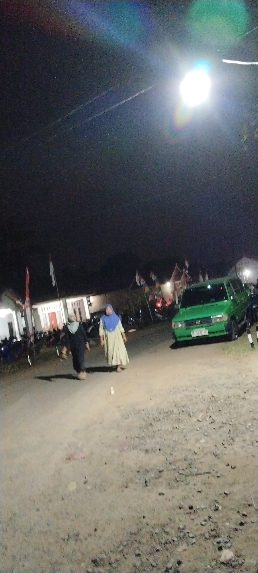 In the city of Sido Rahayu, Indonesia, at night, a dirt road is visible with a green van parked on the right side. Two individuals wearing hijabs are walking on the road. One person on the left is wearing a black abaya, and the person on the right is wearing a beige dress. In the background, there are buildings and flags displayed. The sky is dark with a bright light source in the upper right corner, creating a lens flare effect.