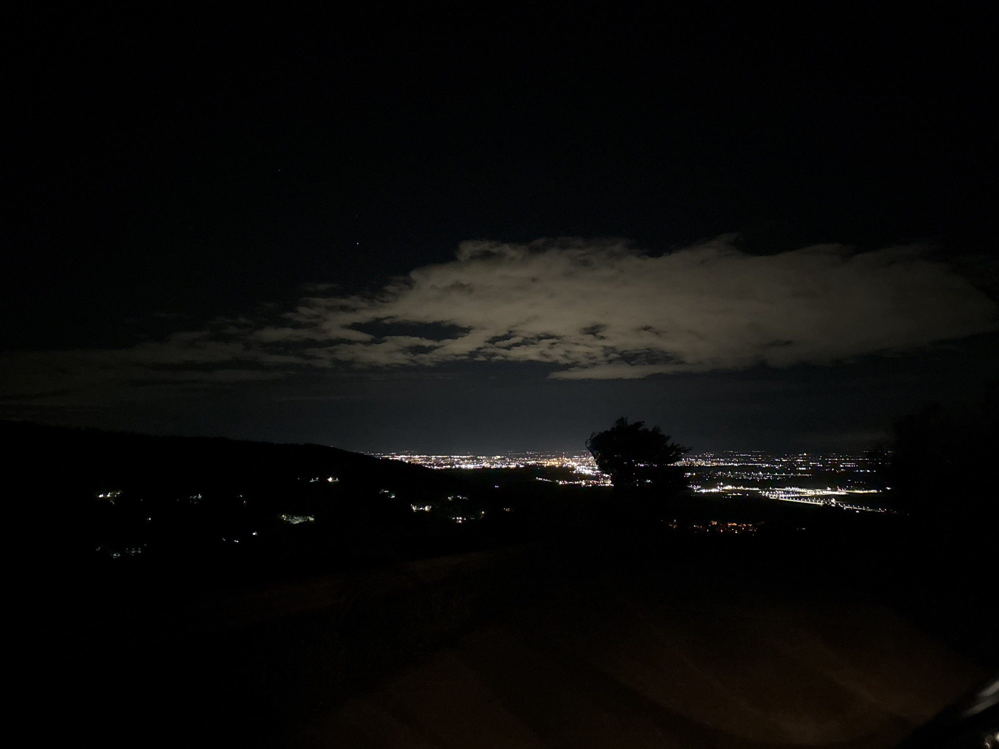The image shows a nighttime view of a city from an elevated vantage point. The city lights are visible in the distance, creating a bright horizon. A large cloud partially obscures the night sky. The foreground is dark, with silhouettes of trees and hills. The location may be Asenovgrad, Bulgaria, but this cannot be confirmed from the image alone.