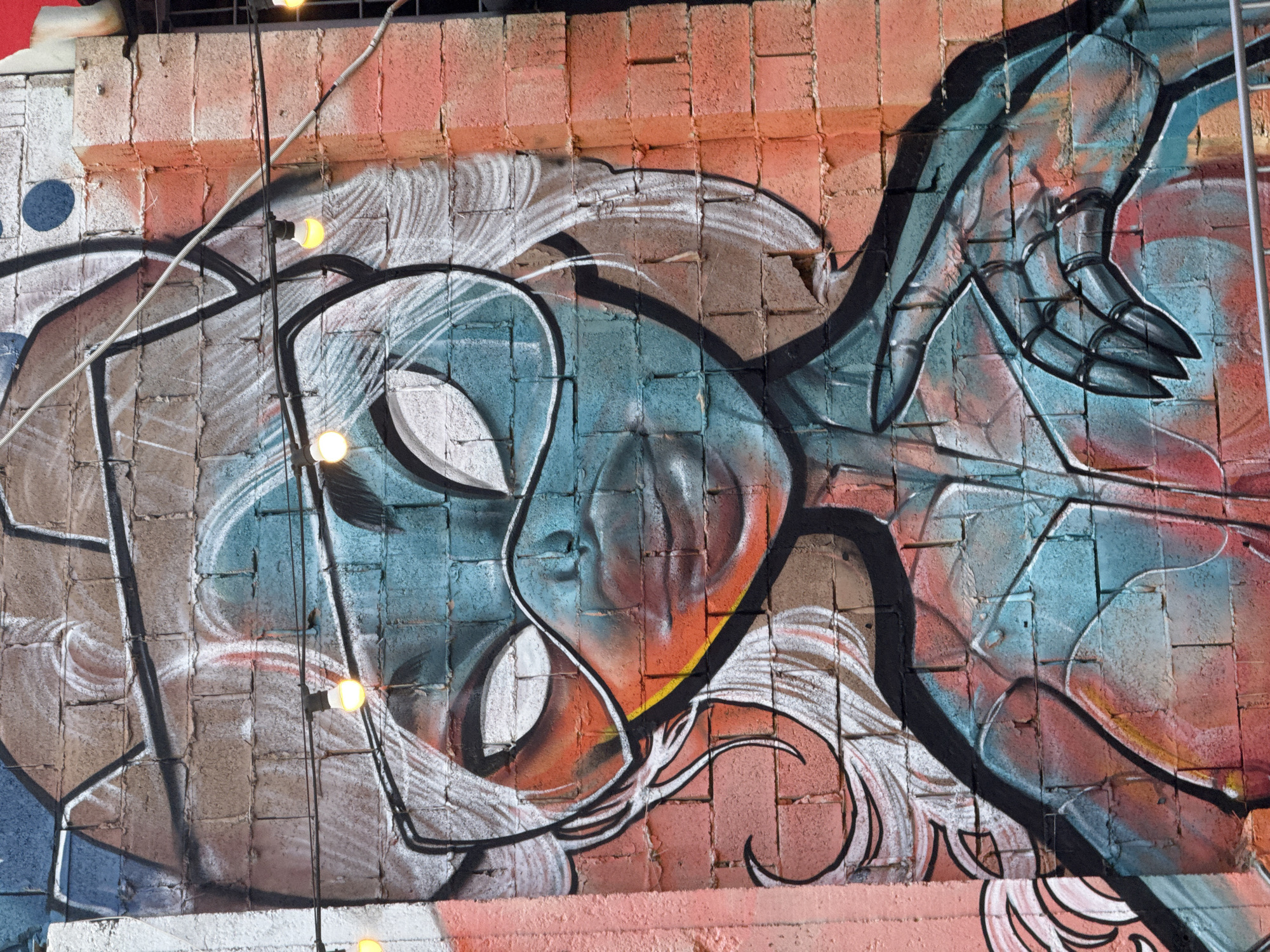 A graffiti artwork of a stylized female figure with blue and red tones is spray-painted on a brick wall in Krasnodar, Russia. The figure has white hair, large white eyes with black outlines, and is wearing a futuristic helmet and goggles. The artwork extends across multiple brick blocks, with some of the paint showing signs of wear and tear on the wall surface. String lights with yellow bulbs are hung across the wall, partially obscuring the graffiti. The background on the right side of the image is a salmon-colored brick wall, while the left side features a pinkish vertical stripe.