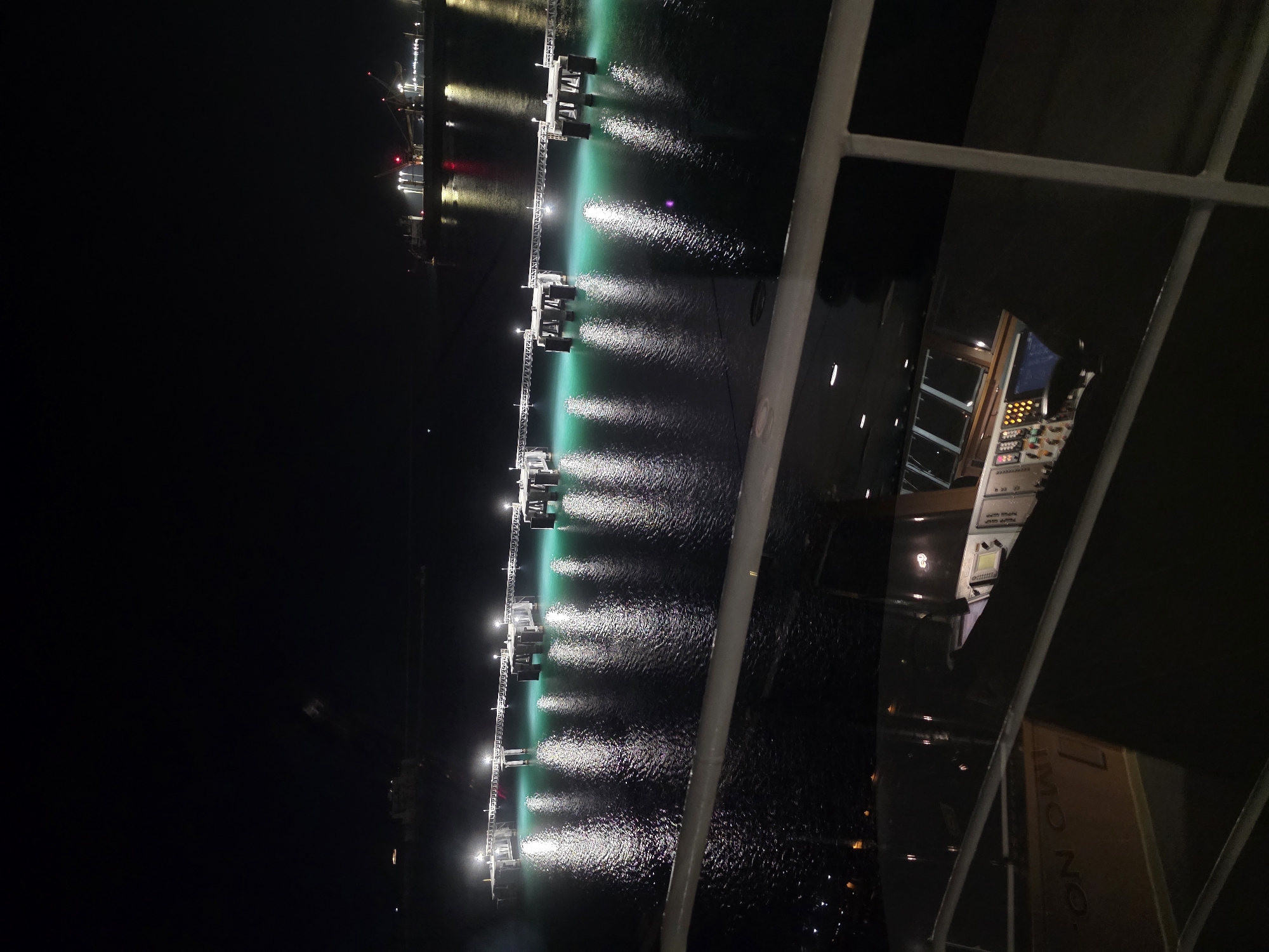 The image depicts a nighttime view from a vessel, looking towards a brightly lit pier extending into dark water. The pier, supported by numerous pilings, is illuminated by a series of lights, casting a greenish glow on the water below. Reflections of these lights shimmer on the rippling surface. In the background, distant lights suggest industrial activity or a shoreline. The foreground shows a railing and a glimpse of a vessel's interior, possibly a bridge or control room, with illuminated panels and screens visible through a reflective surface. The scene is set at night in the port of Brindisi, Italy, looking out towards the Adriatic Sea.