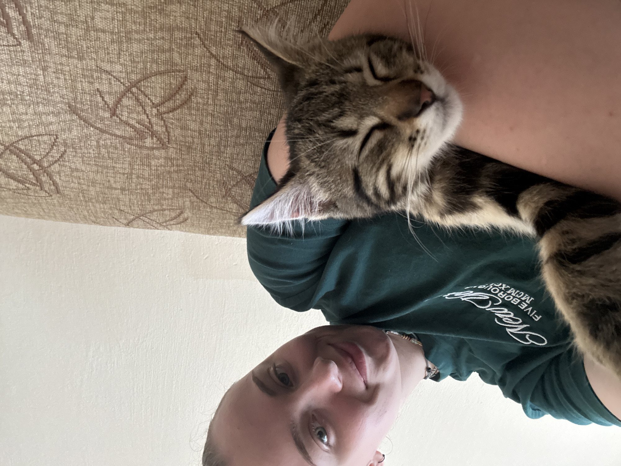 A young woman with blonde hair pulled back is taking a selfie. She is wearing a dark green t-shirt with white writing on it. She is smiling and looking at the camera. Her left arm is visible, and a tabby cat is resting on it, facing away from the camera. The cat's eyes are closed, and it appears to be sleeping. The woman is holding the cat with her left arm. They are indoors, as indicated by the wall in the background. A textured couch is visible to the right. The overall impression is one of comfort and affection. There is no information available about the location in the city of Gabrovo, Bulgaria.