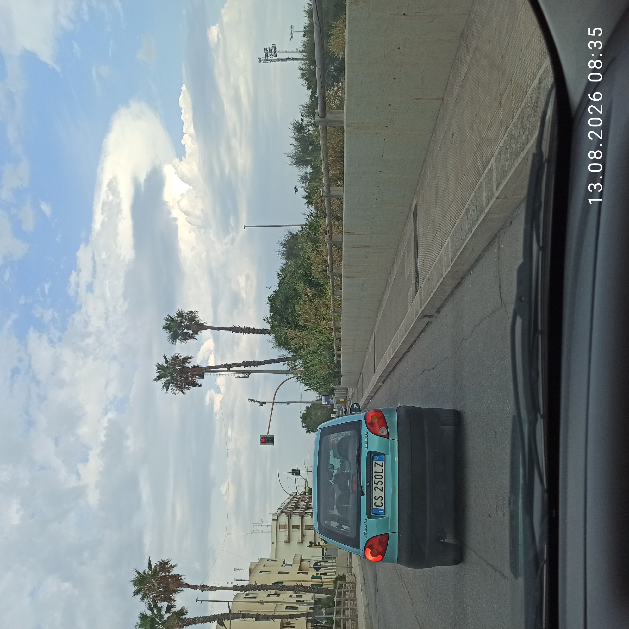 The image shows a street scene in Molfetta, Italy. In the foreground, the dashboard and windshield of a vehicle are visible, suggesting the photo was taken from inside a car. A blue Daewoo Matiz car with the license plate "CS 250LZ" is traveling ahead on a paved road. The car's brake lights are illuminated. To the right of the road, there is a concrete wall with a wooden railing on top, bordering a sidewalk with textured paving. Beyond the wall, there are trees and foliage, and further in the background, there are buildings, streetlights, and palm trees lining the street. The sky is cloudy with patches of blue. A traffic light with a red signal is visible on the right side of the road. The timestamp "13.08.2026 08:35" is overlaid on the image.