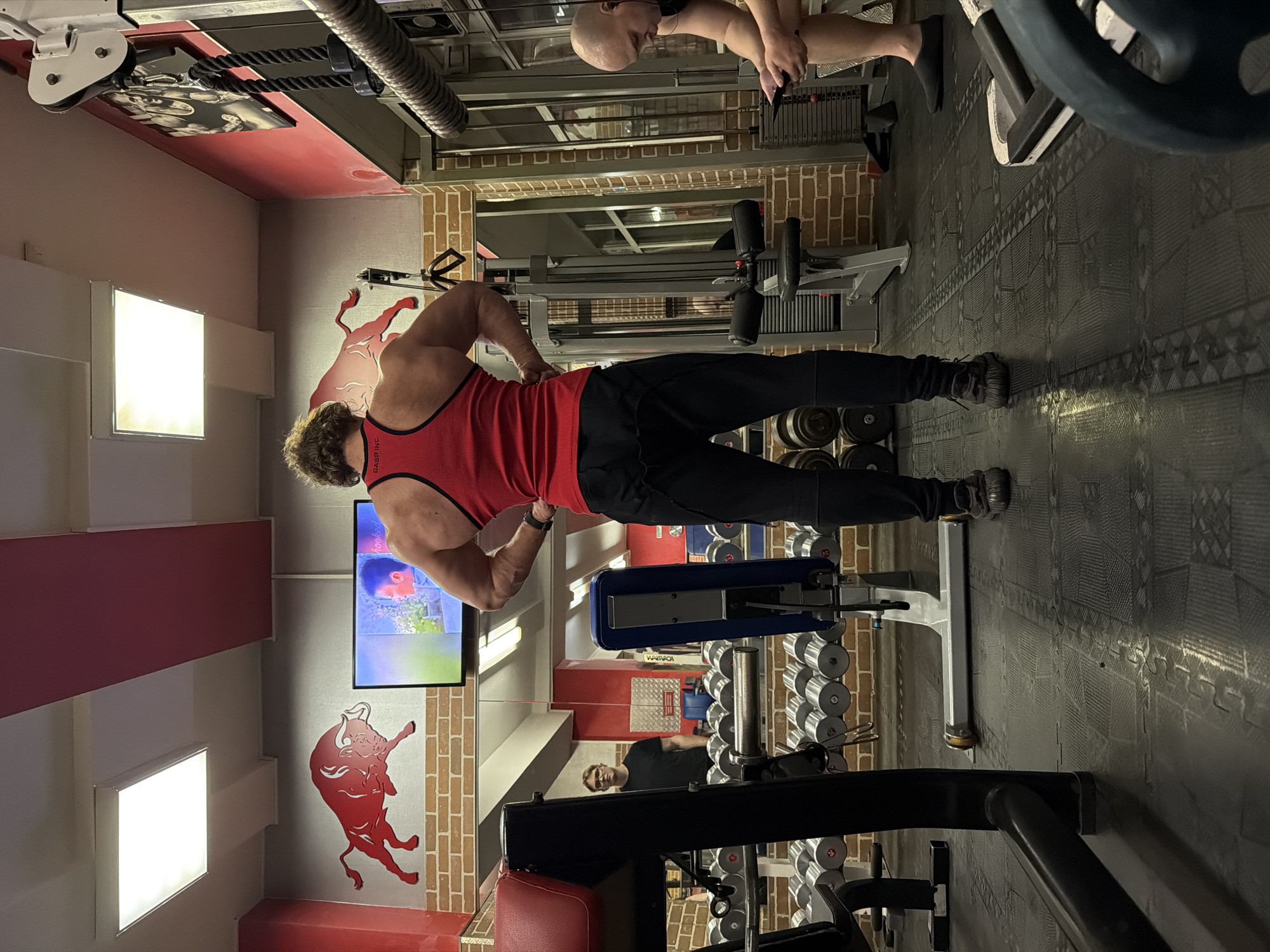 An interior view captures a gymnasium in Saint Petersburg, Russia, characterized by brick-patterned lower walls and grey upper walls accented with red stripes and two red bull decals. The primary subject, a muscular male wearing a red tank top with "GASP INC" branding and black athletic pants, stands with his back to the viewer, hands on his hips, displaying his developed back musculature. To the left, a large mirror reflects another male individual in a black t-shirt and glasses, standing near a rack of numerous silver dumbbells. To the right, a bald male subject in a black t-shirt and shorts sits on a bench, intently focused on a mobile phone in his hands. Various gym equipment, including cable machines and weight benches, are visible throughout the space. A flat-screen television is mounted on the wall above the mirror, displaying an image of a person. The floor is covered with black interlocking rubber mats. Overhead, rectangular fluorescent light panels illuminate the scene.